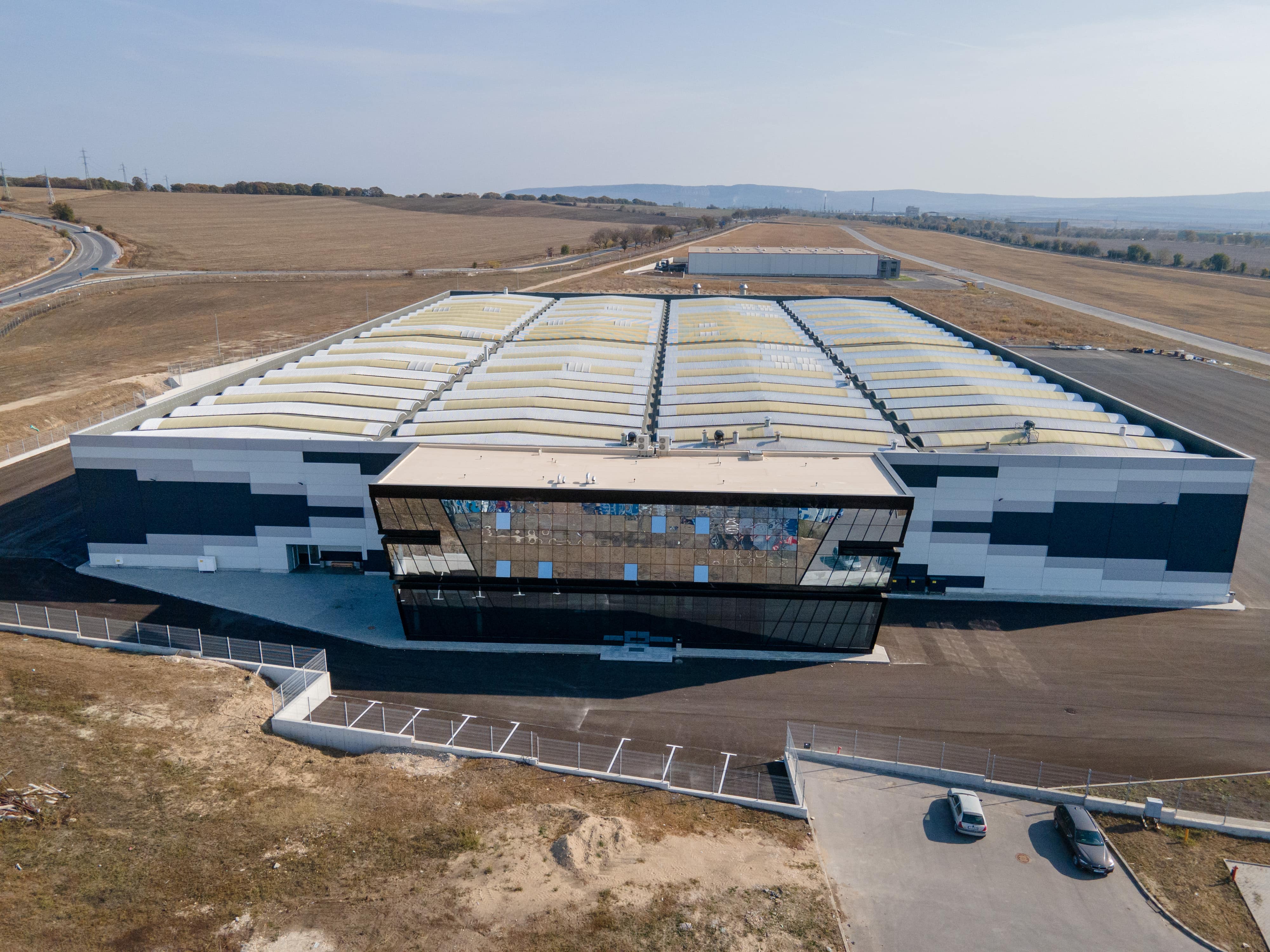 Aerial view of a large industrial building with barrel-vault roof and glazed entrance facade in a flat arid landscape