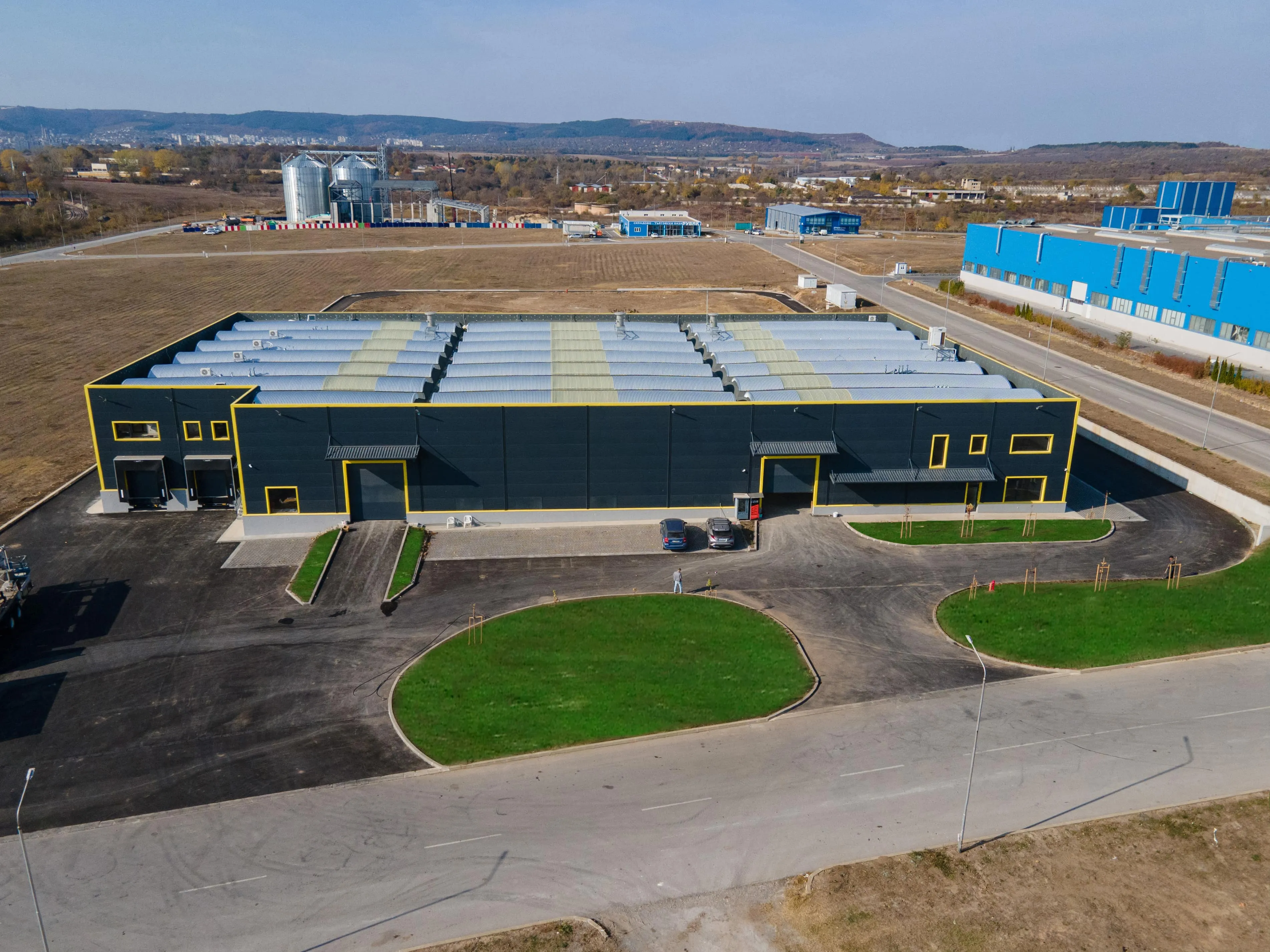 Aerial view of a large dark industrial warehouse with yellow accents and barrel-vault roof in an open rural area