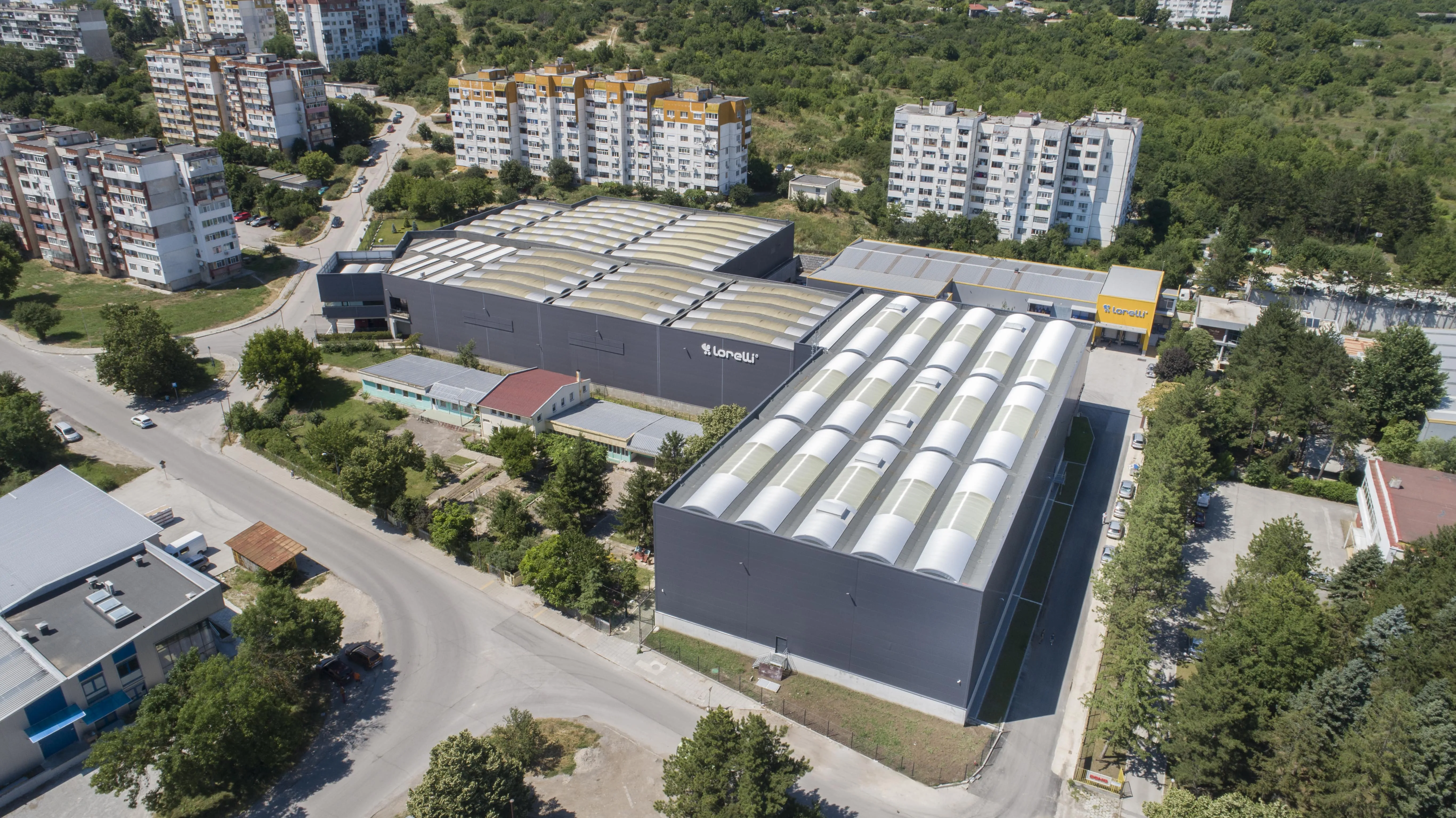 Aerial view of dark grey industrial warehouses with barrel-vault roofs bearing Lonelli branding, urban surroundings