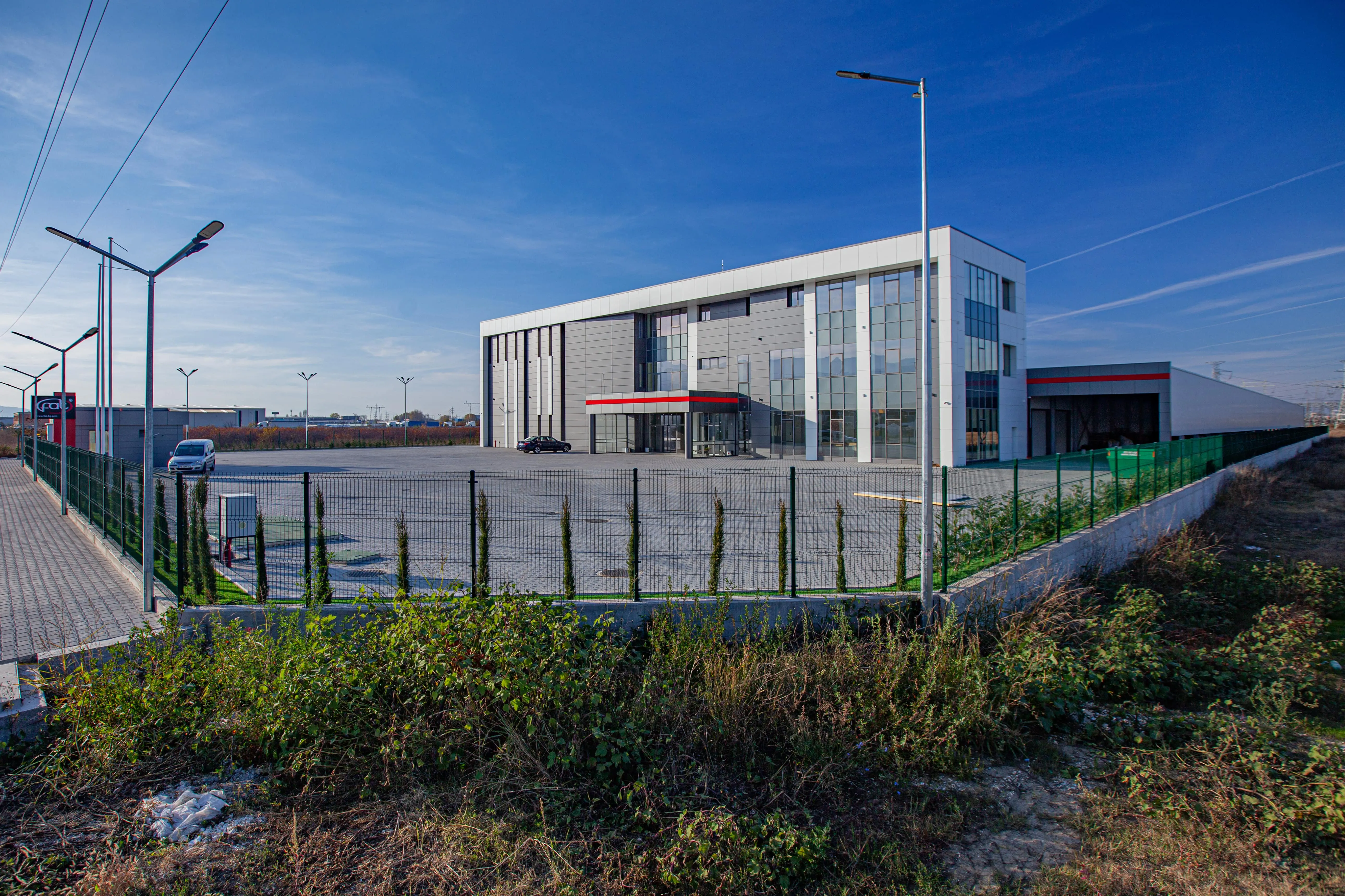 Modern two-storey commercial building with white and grey panels and red entrance canopy, fenced industrial yard
