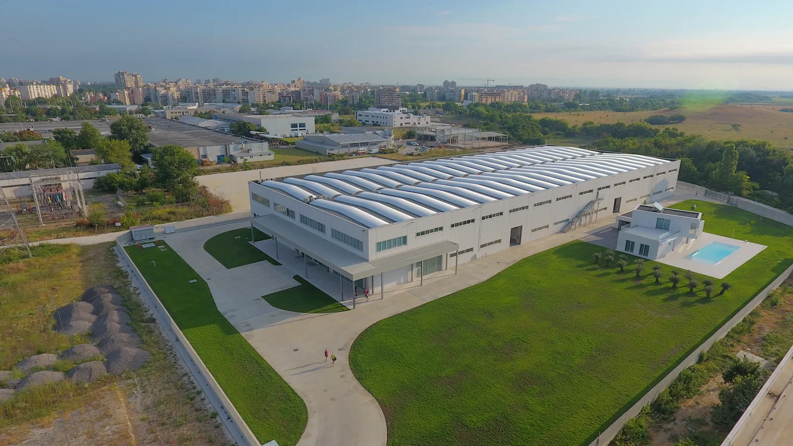 Aerial view of a large white industrial warehouse with barrel-vault roof skylights, green lawn, and outdoor pool