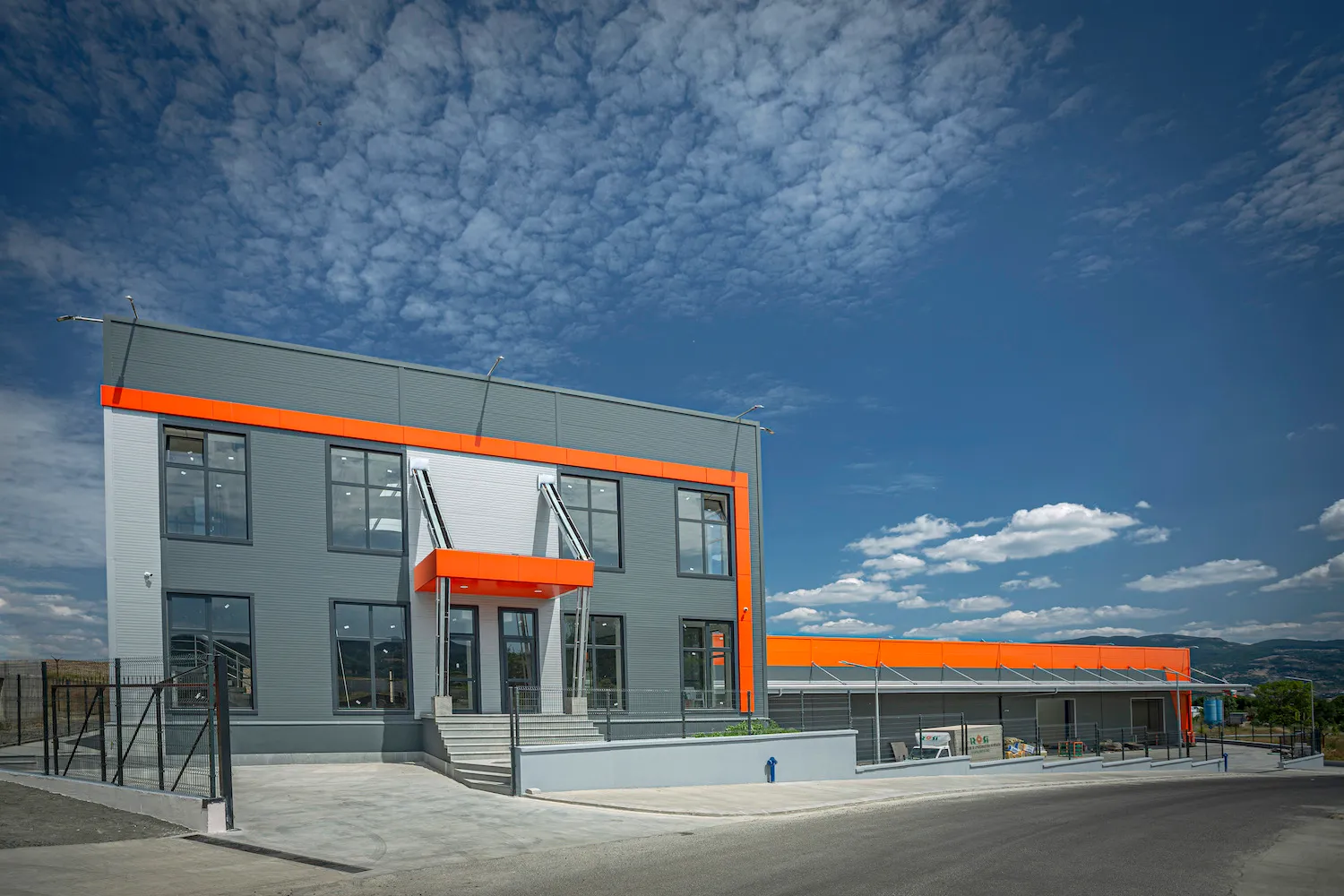 Two-storey industrial office building with grey and orange cladding against a blue sky with scattered clouds