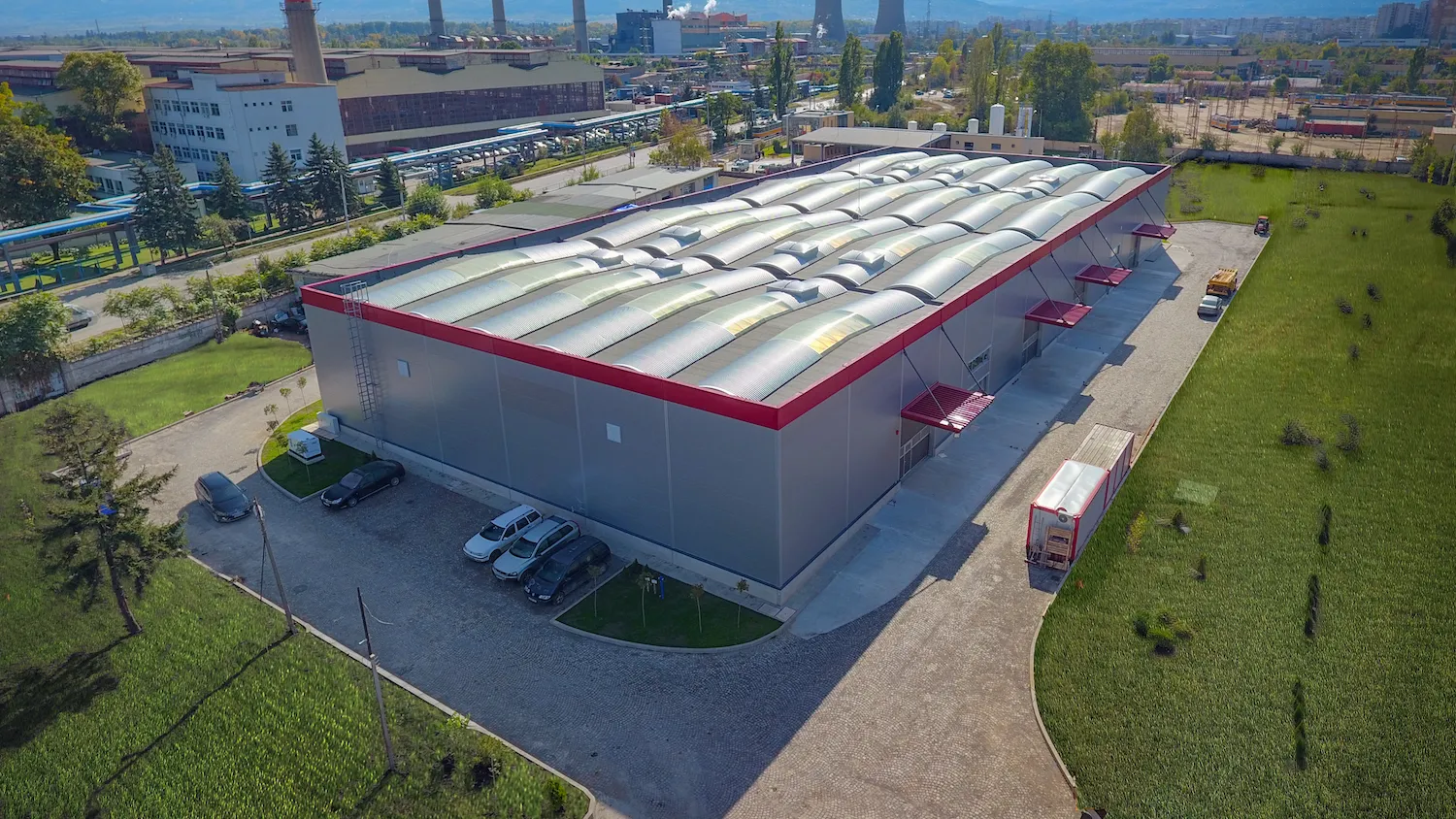 Aerial view of gray industrial warehouse with red trim accents and barrel-vault roof near industrial area with chimneys
