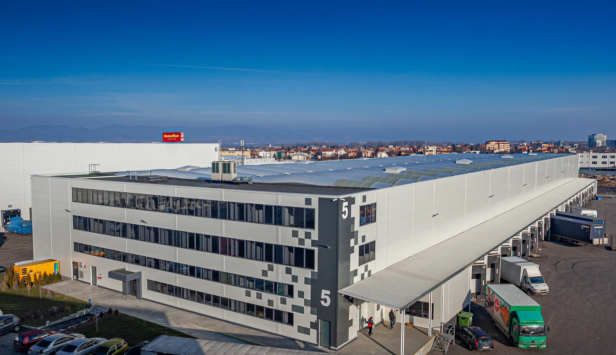 Aerial view of logistics warehouse building number 5 with checkered office facade, loading docks and delivery trucks