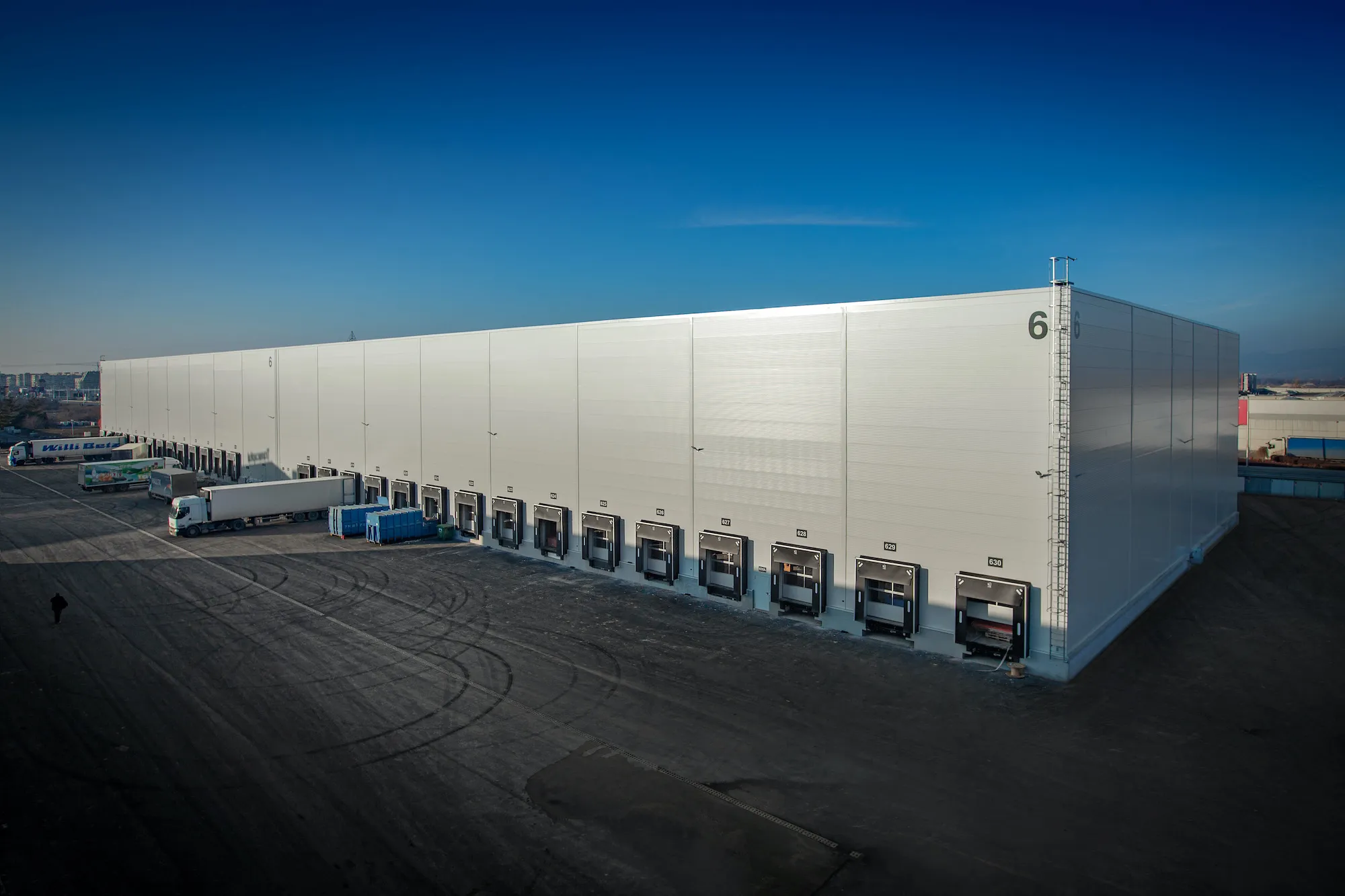 Exterior facade of large white logistics warehouse building number 6 with multiple loading docks and trucks alongside
