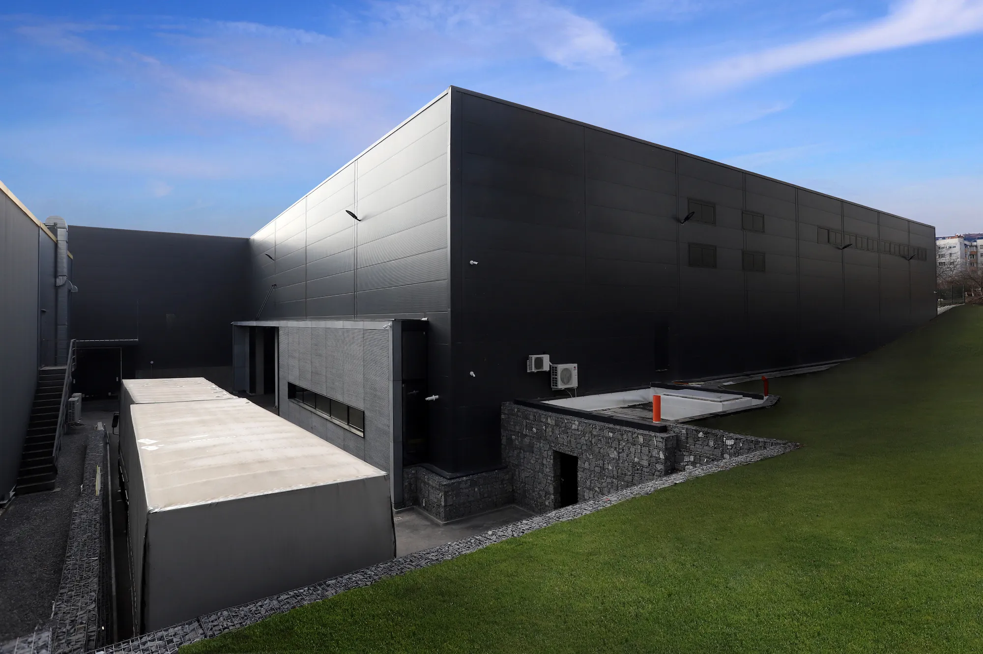 Large dark charcoal-clad industrial building with stone gabion wall feature, loading ramp, and green lawn