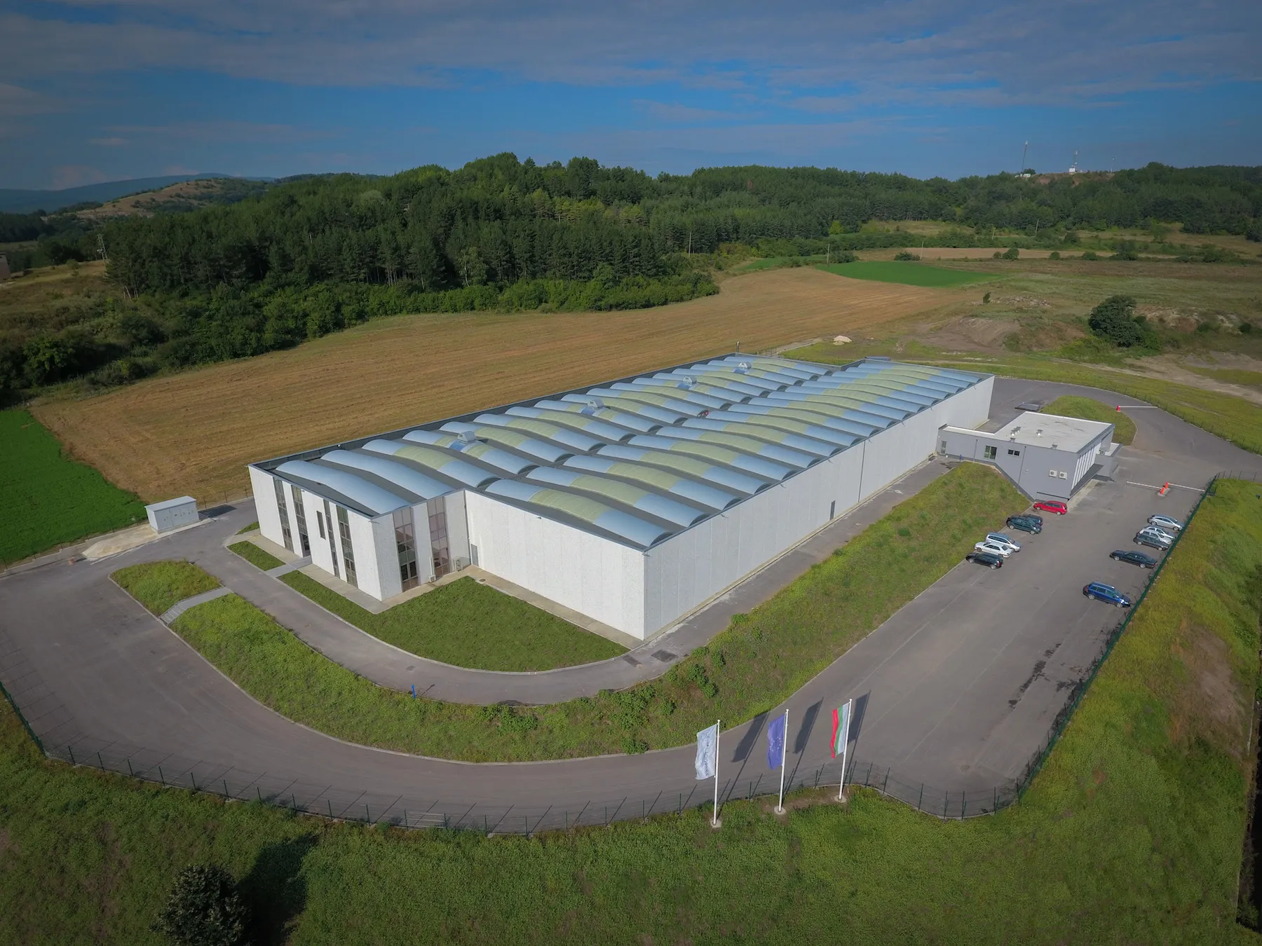 Aerial view of white single-story industrial warehouse surrounded by green fields with office annex and parking area