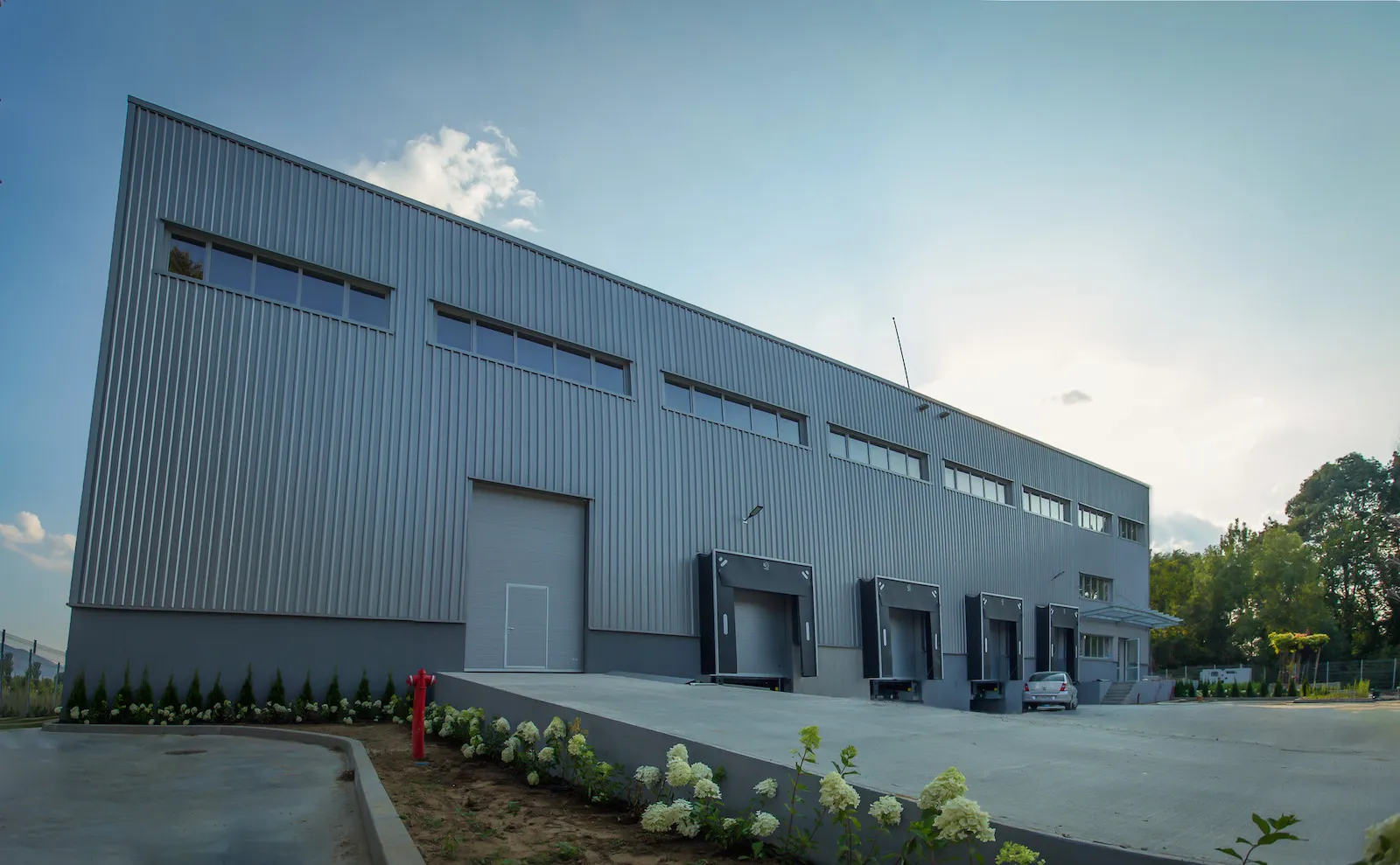 Gray metal-clad warehouse building with multiple loading dock doors and white flowering shrubs along the facade