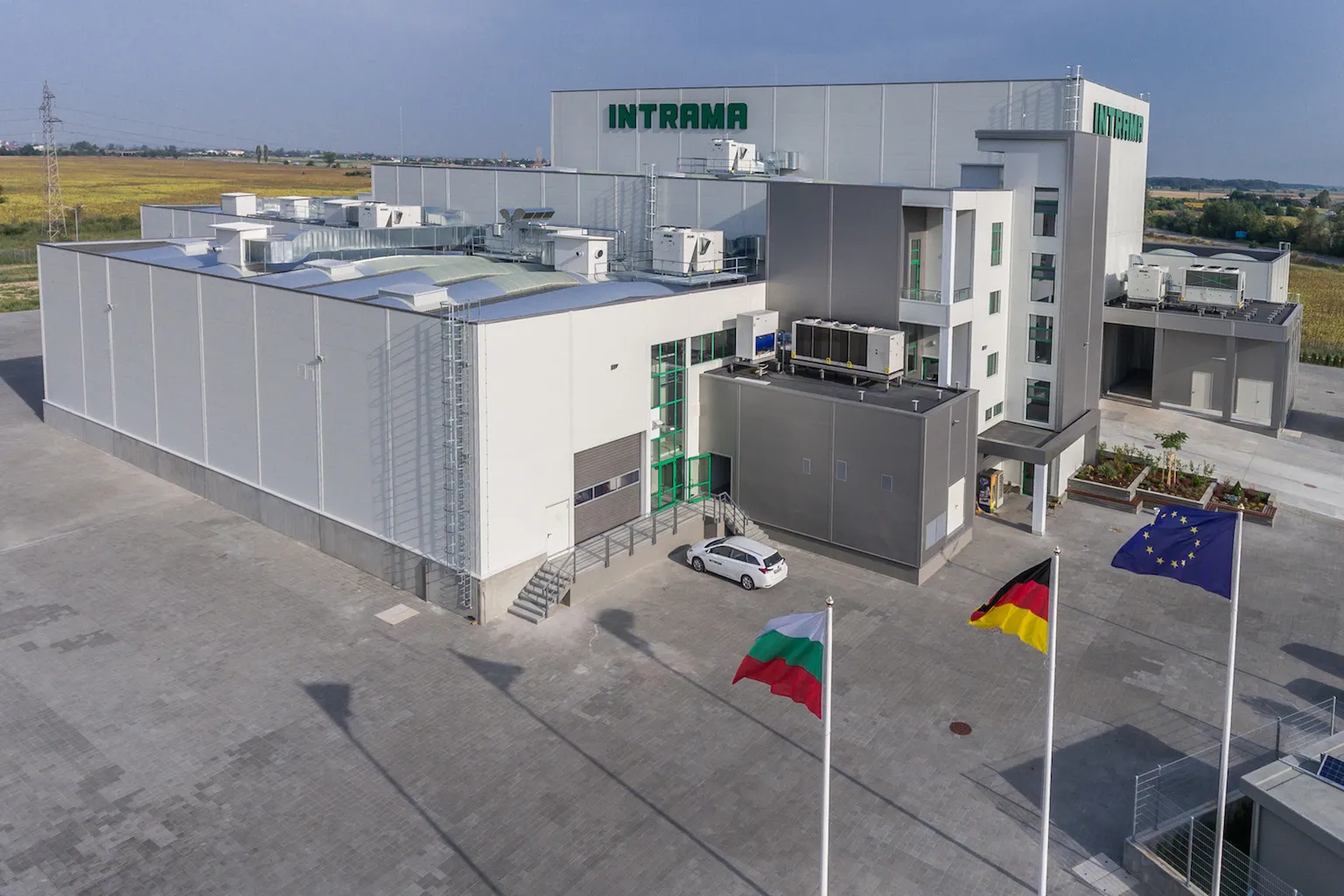 Aerial view of Intrama industrial facility with white warehouse and multi-story office block, EU flags in foreground
