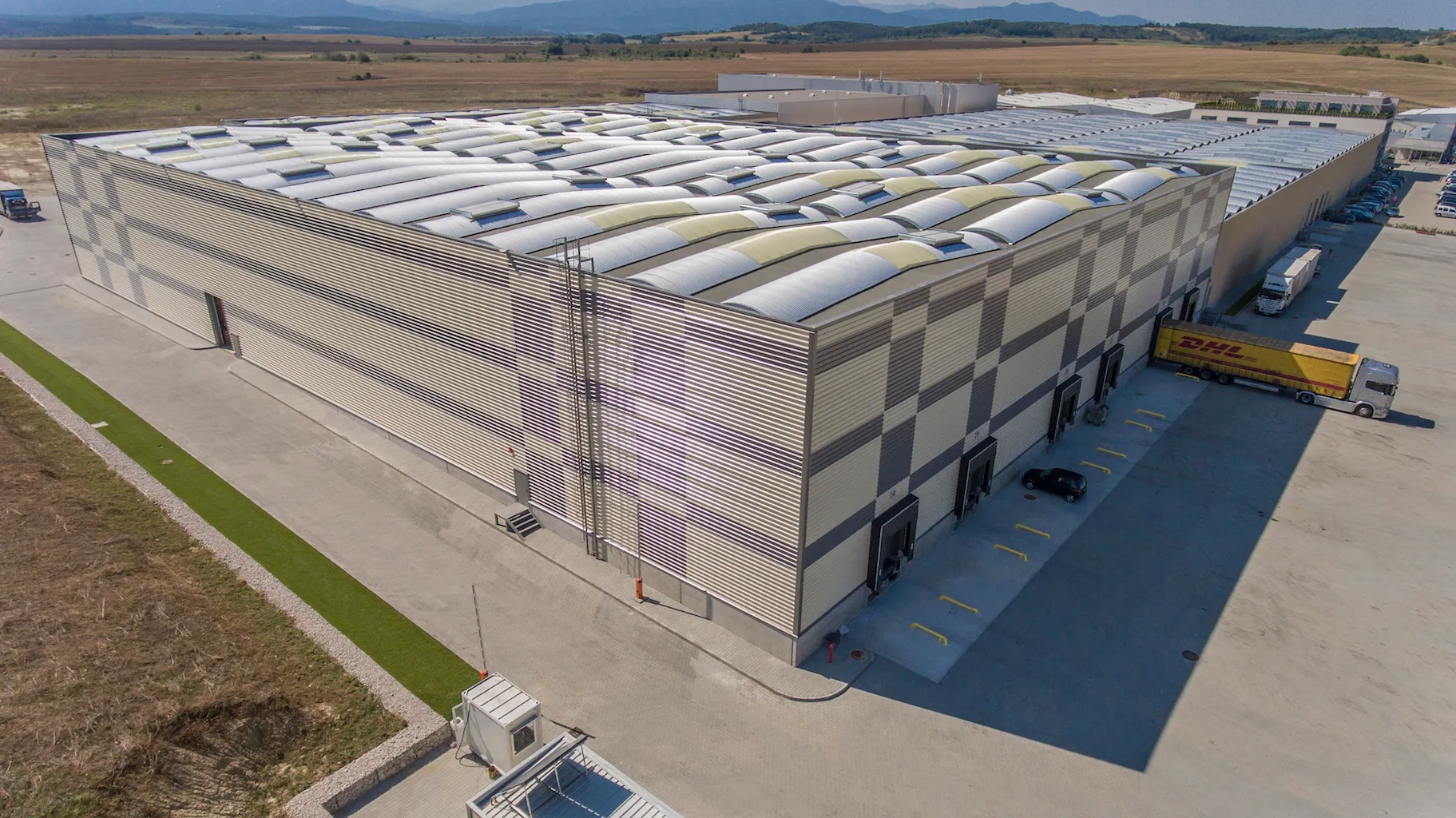 Aerial view of large industrial warehouse with corrugated roof and checkered cladding, DHL truck at loading dock