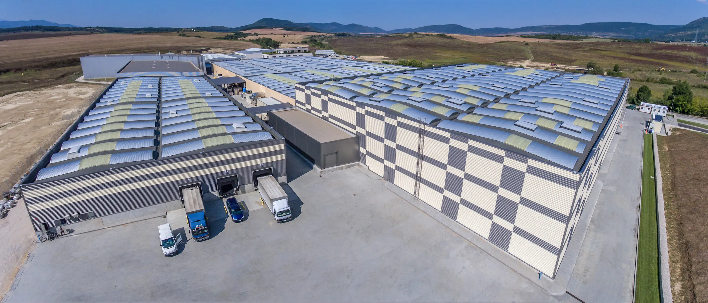 Aerial view of large industrial warehouse with checkered facade pattern and multiple loading docks, trucks in foreground