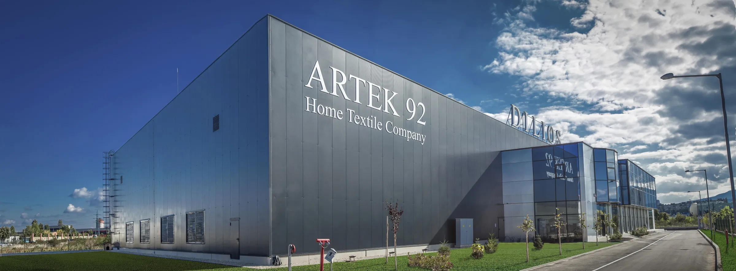 Large dark grey Artek 92 Home Textile Company industrial building with glass showroom wing under blue sky