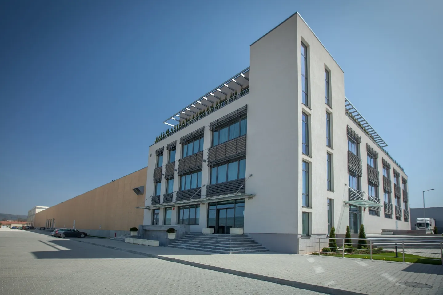 Four-story modern office building with white render, horizontal louvre shading, rooftop terrace, and warehouse wing