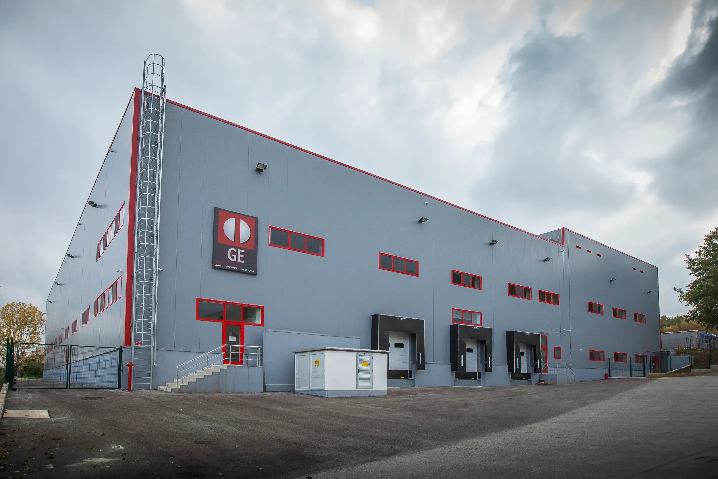 Grey two-story industrial facility with red window frames and GE logo signage, featuring loading docks
