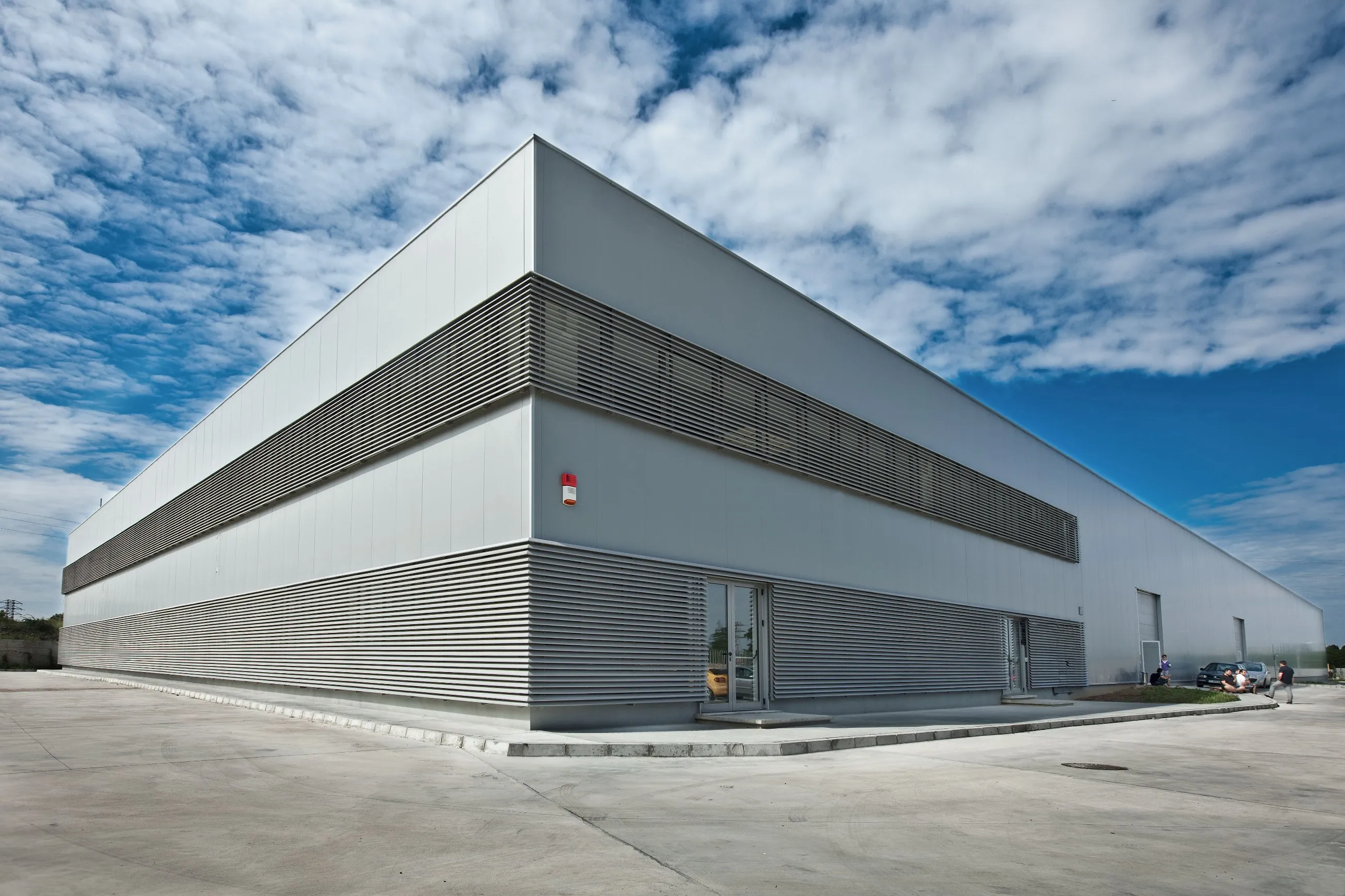 Modern white industrial building with horizontal louvre cladding bands and glass entrance under blue cloudy sky