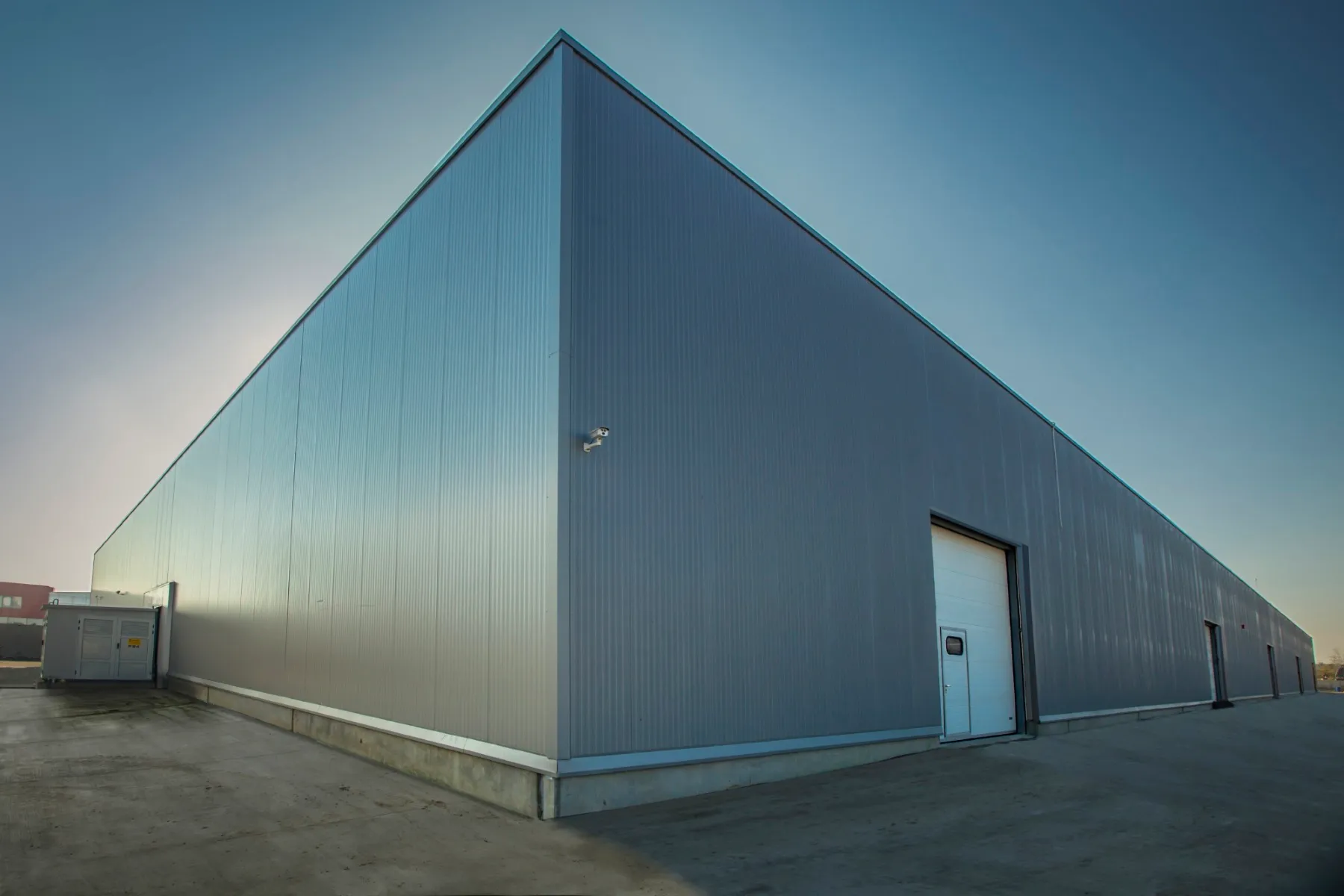 Corner view of a large grey metal-panel warehouse with a white roller door and security camera at dusk