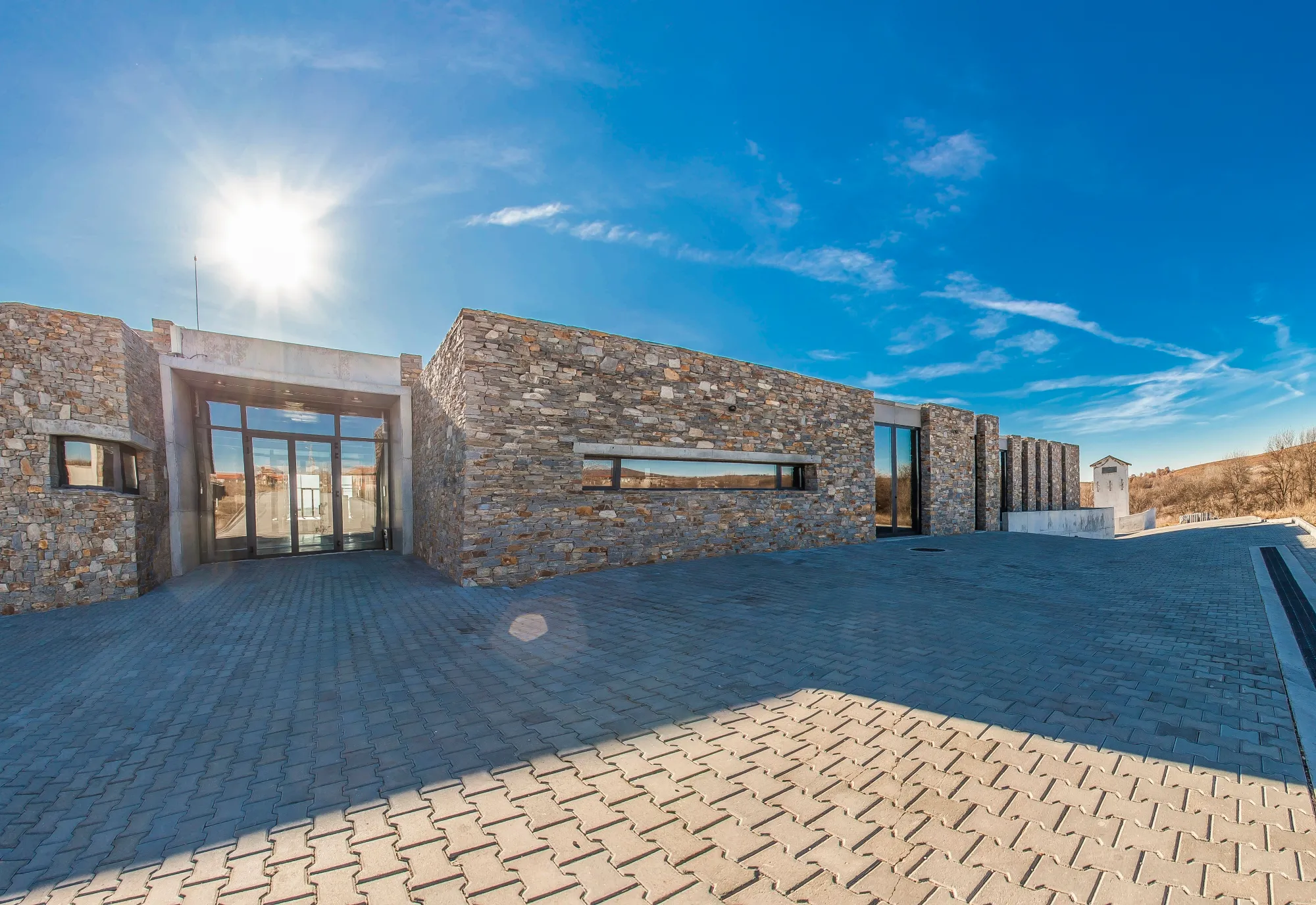 Modern single-story stone-clad winery or estate building with large glass doors and paved courtyard under bright sun