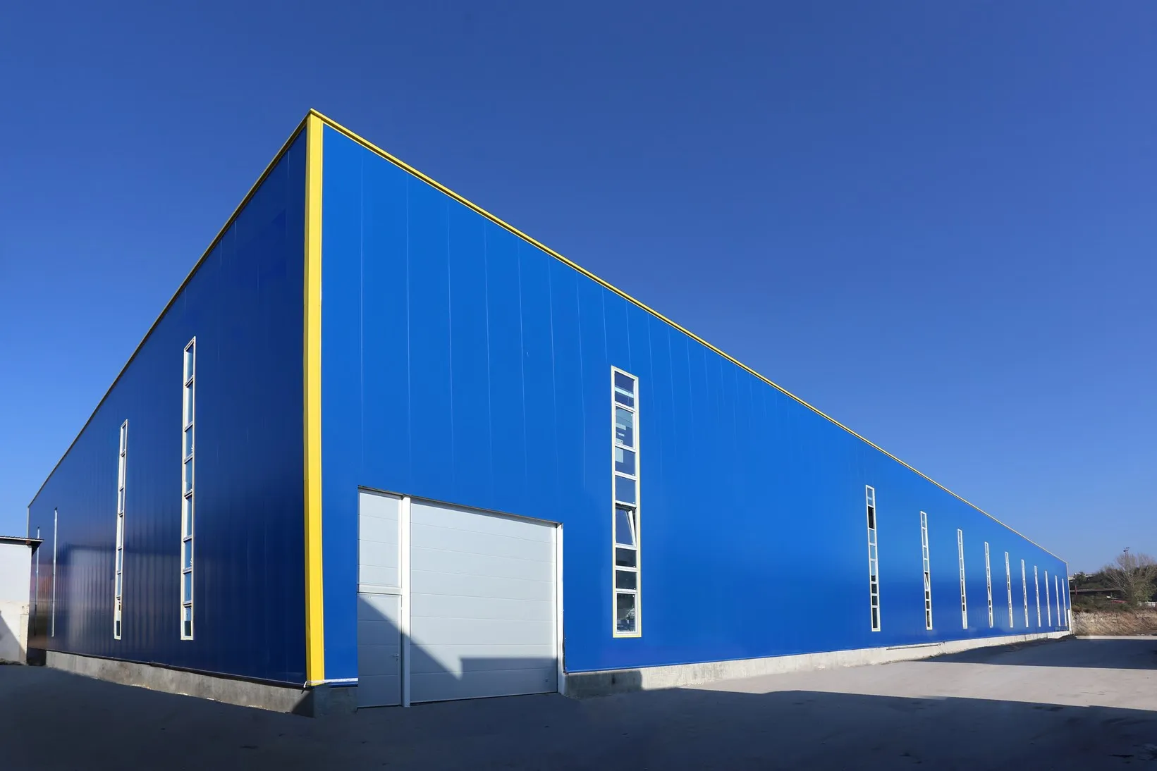 Exterior of a large blue metal warehouse with yellow details and a white roller door under a clear blue sky