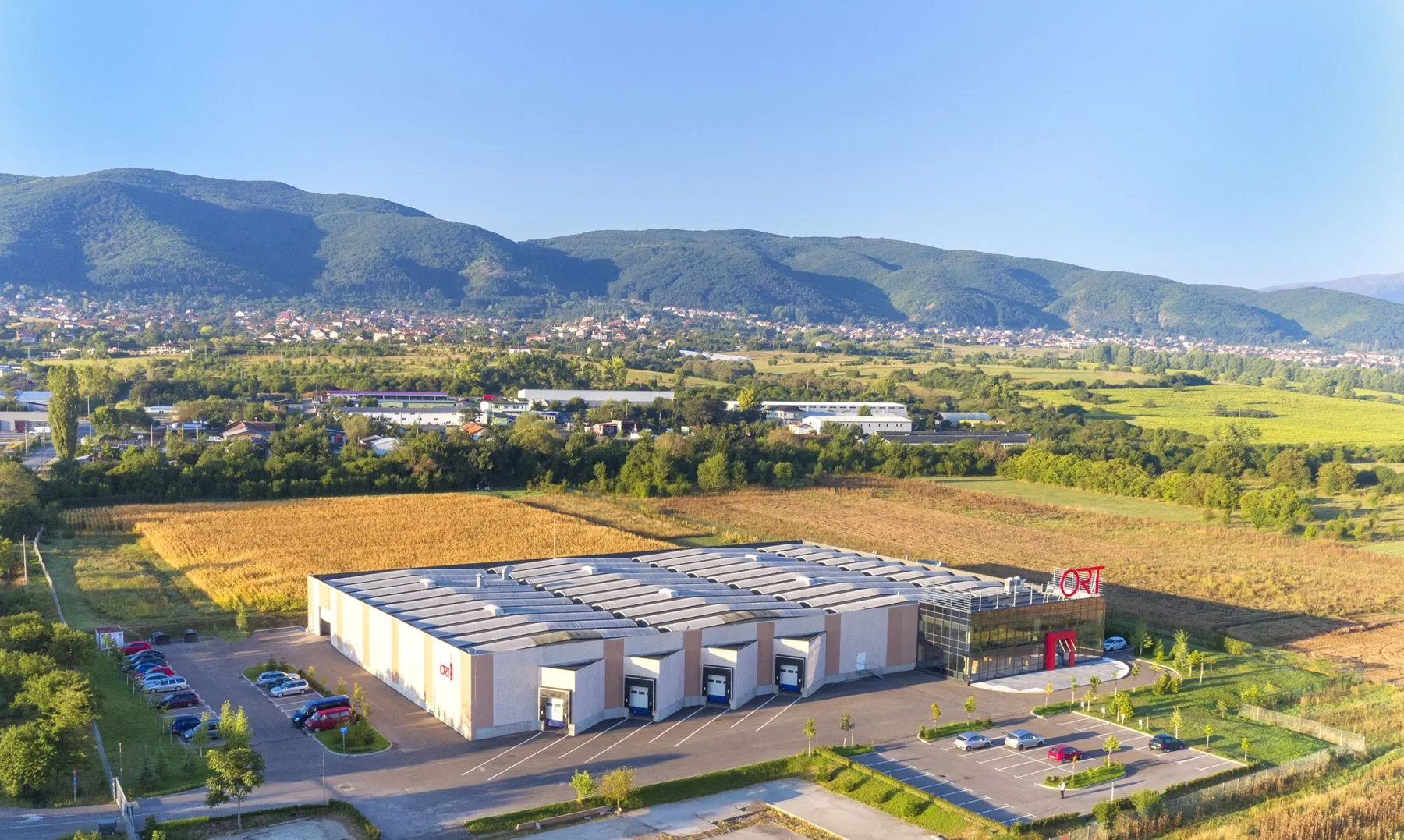 Aerial view of a large ORT-branded industrial complex with solar roof panels set against green hills and farmland