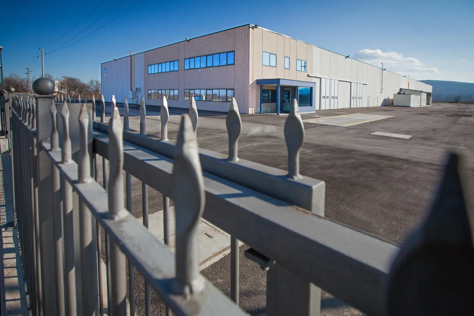 Two-story industrial building with beige facade viewed through a metal security fence on a clear winter day