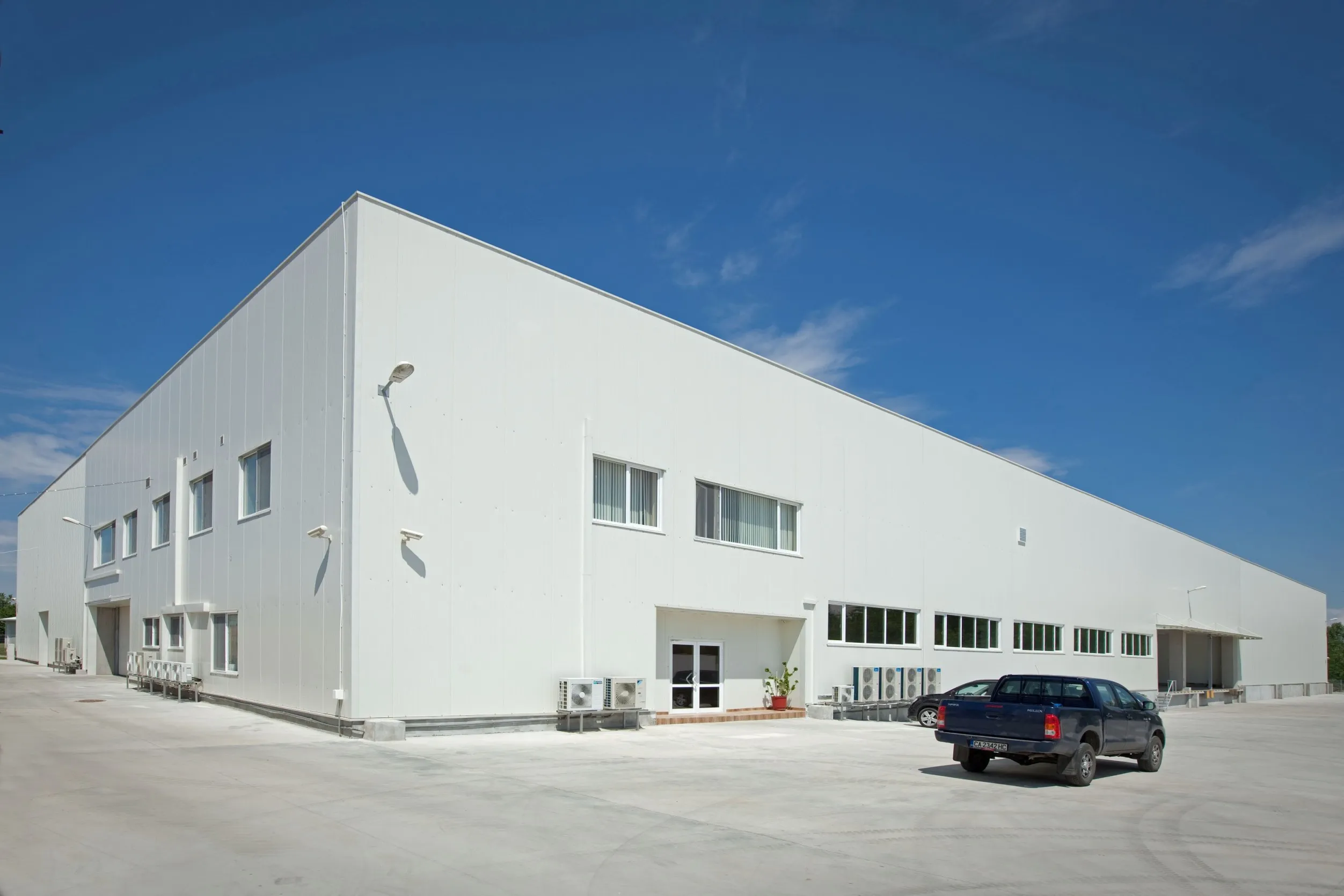 Large white single-storey industrial warehouse with flat facade and a dark blue pickup truck in the foreground
