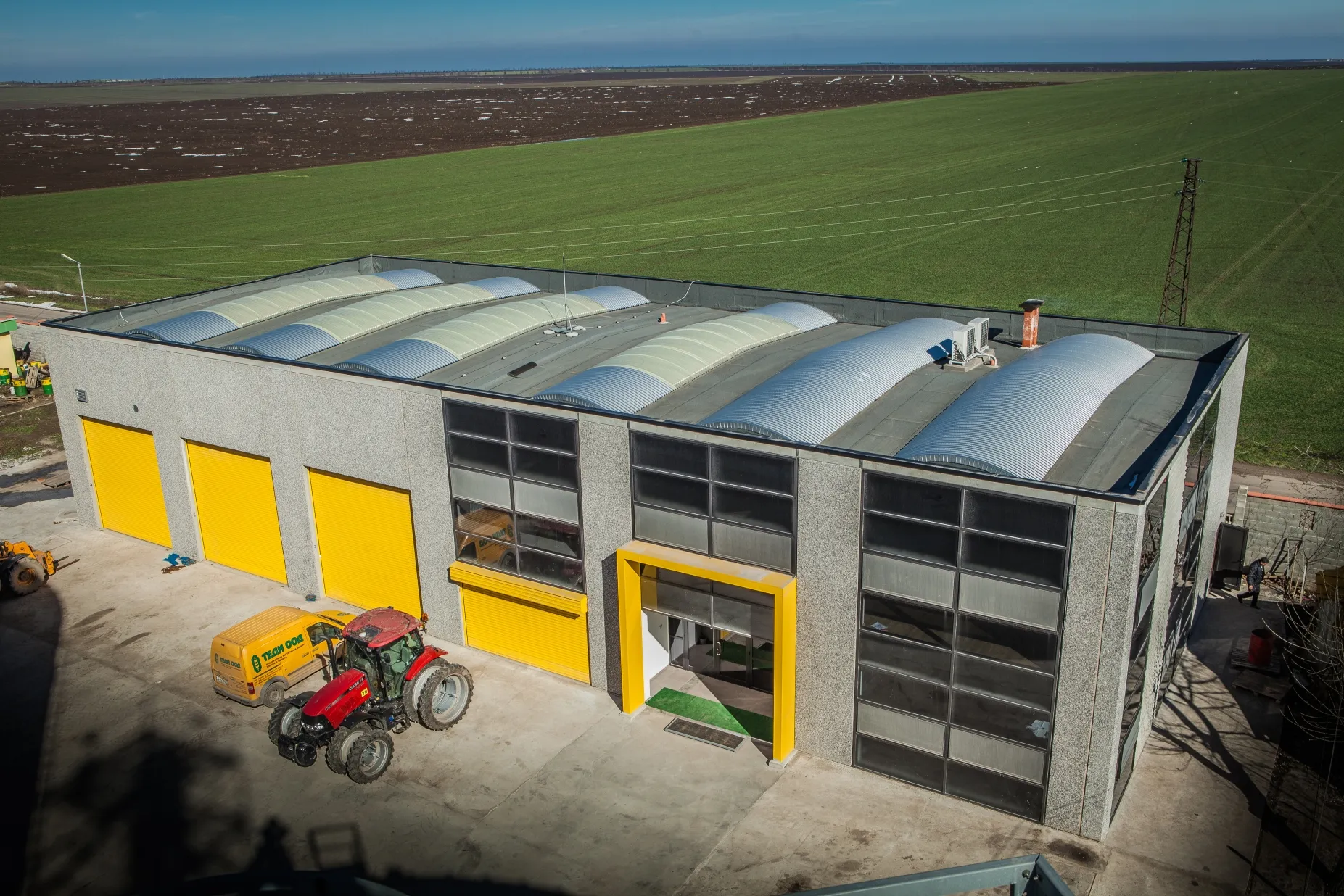 Aerial view of grey agricultural service building with yellow roller shutters, a red tractor parked in front