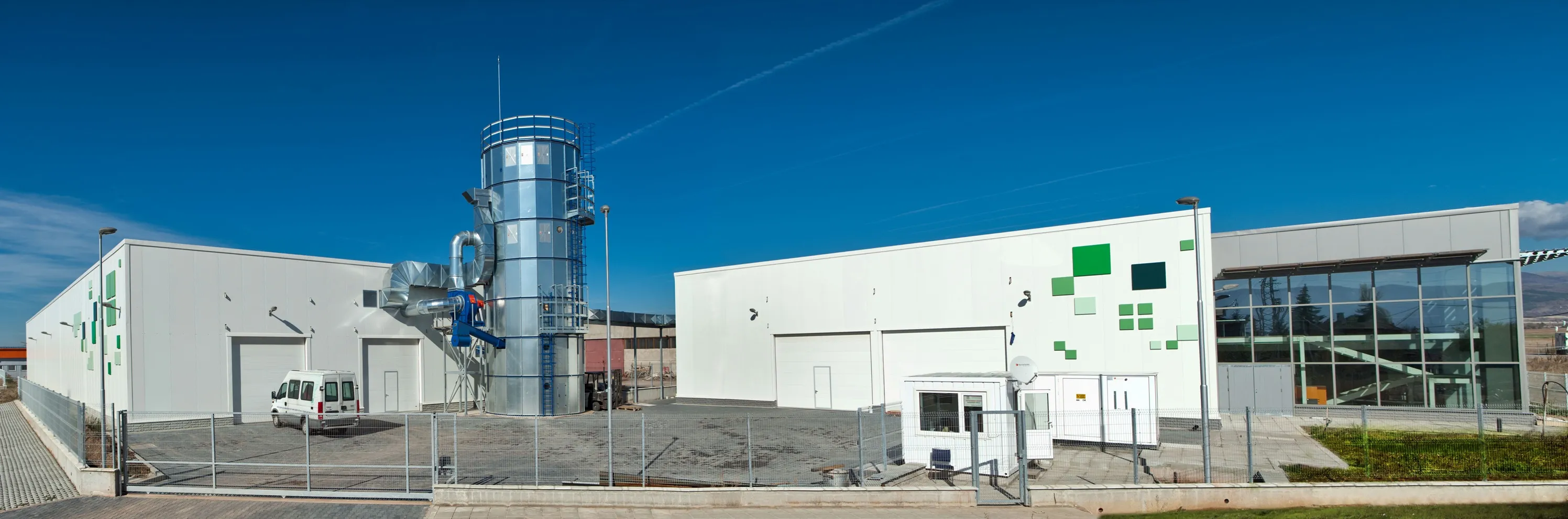 Panoramic view of white industrial facility with a tall metal processing tower and green square decorative accents