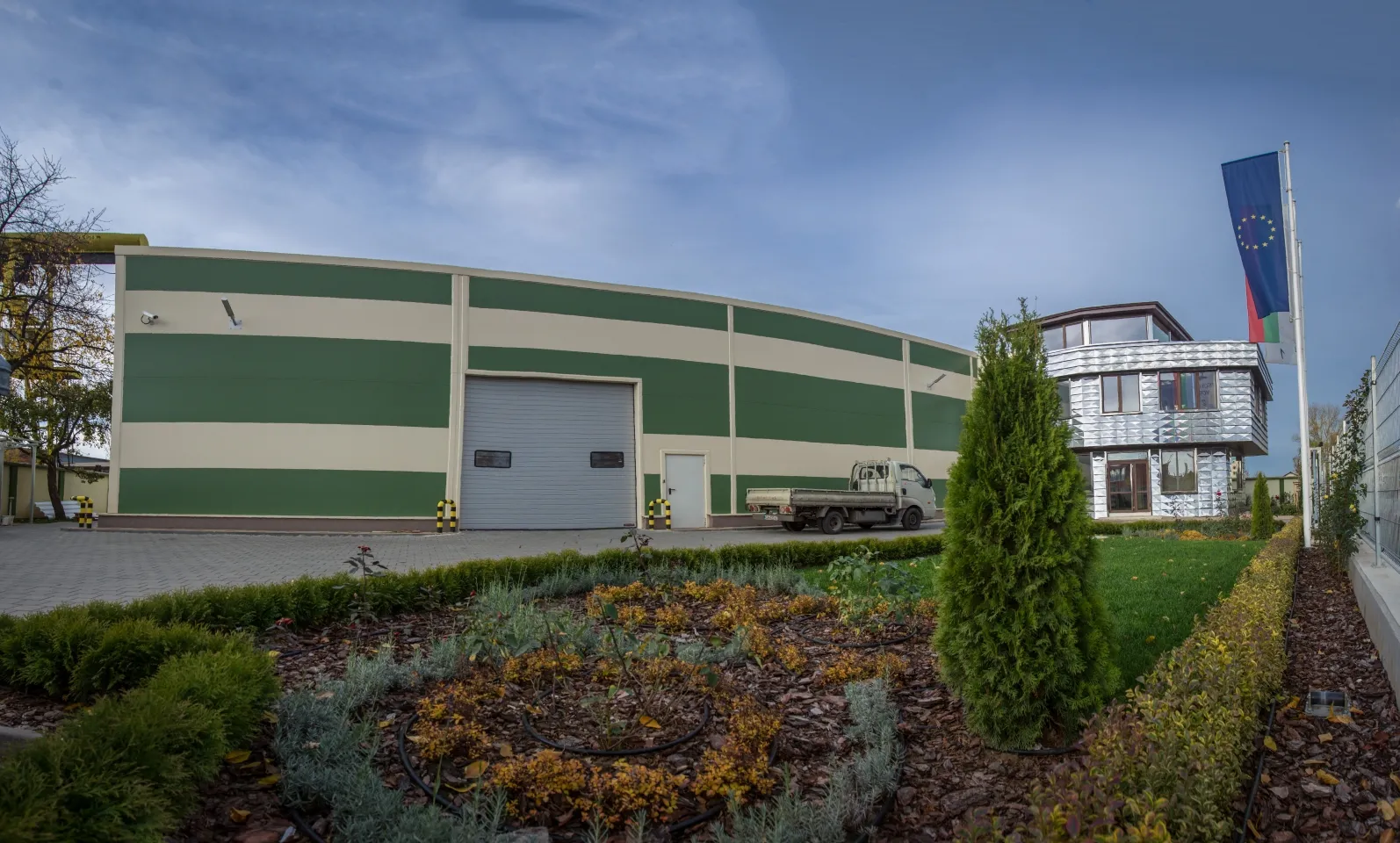Curved green and cream industrial warehouse with roller shutter gate, landscaped garden and EU and Bulgarian flags