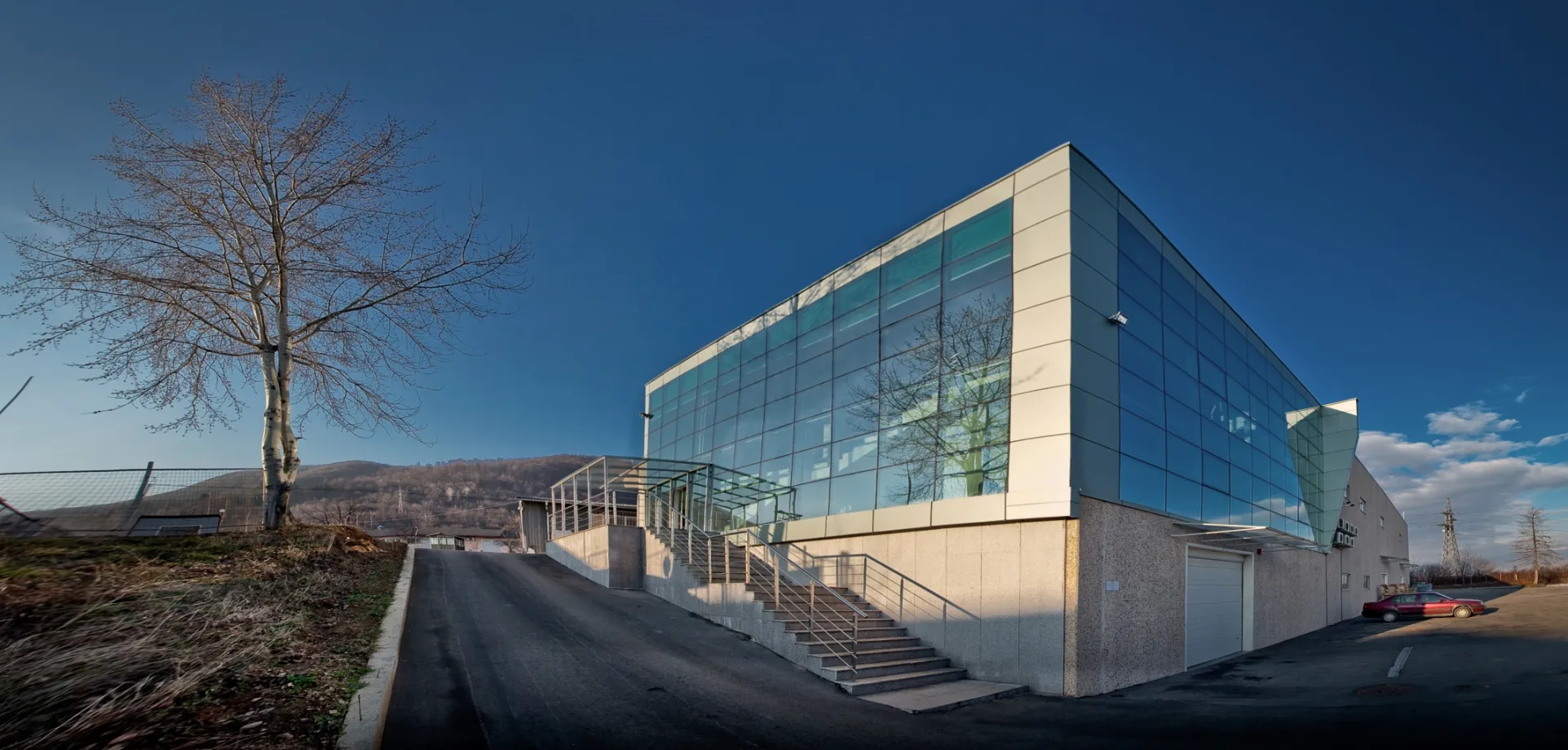 Modern office and production building with full-height curtain glass facade, exterior staircase, and hillside backdrop