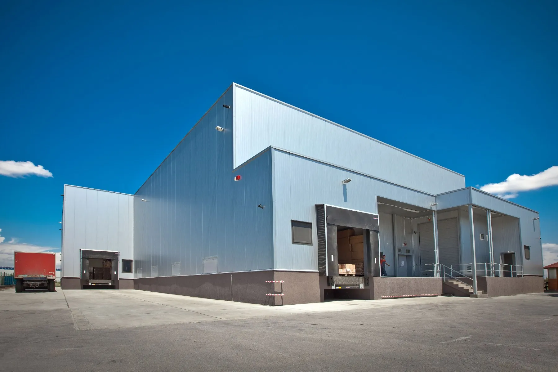 Light blue steel-clad warehouse with multiple loading dock doors, a red truck parked alongside under blue sky