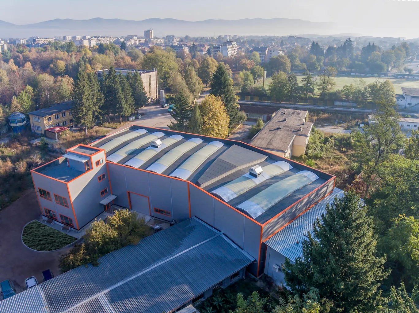 Aerial view of grey industrial complex with orange-trimmed roofline and arched skylights, surrounded by autumn trees