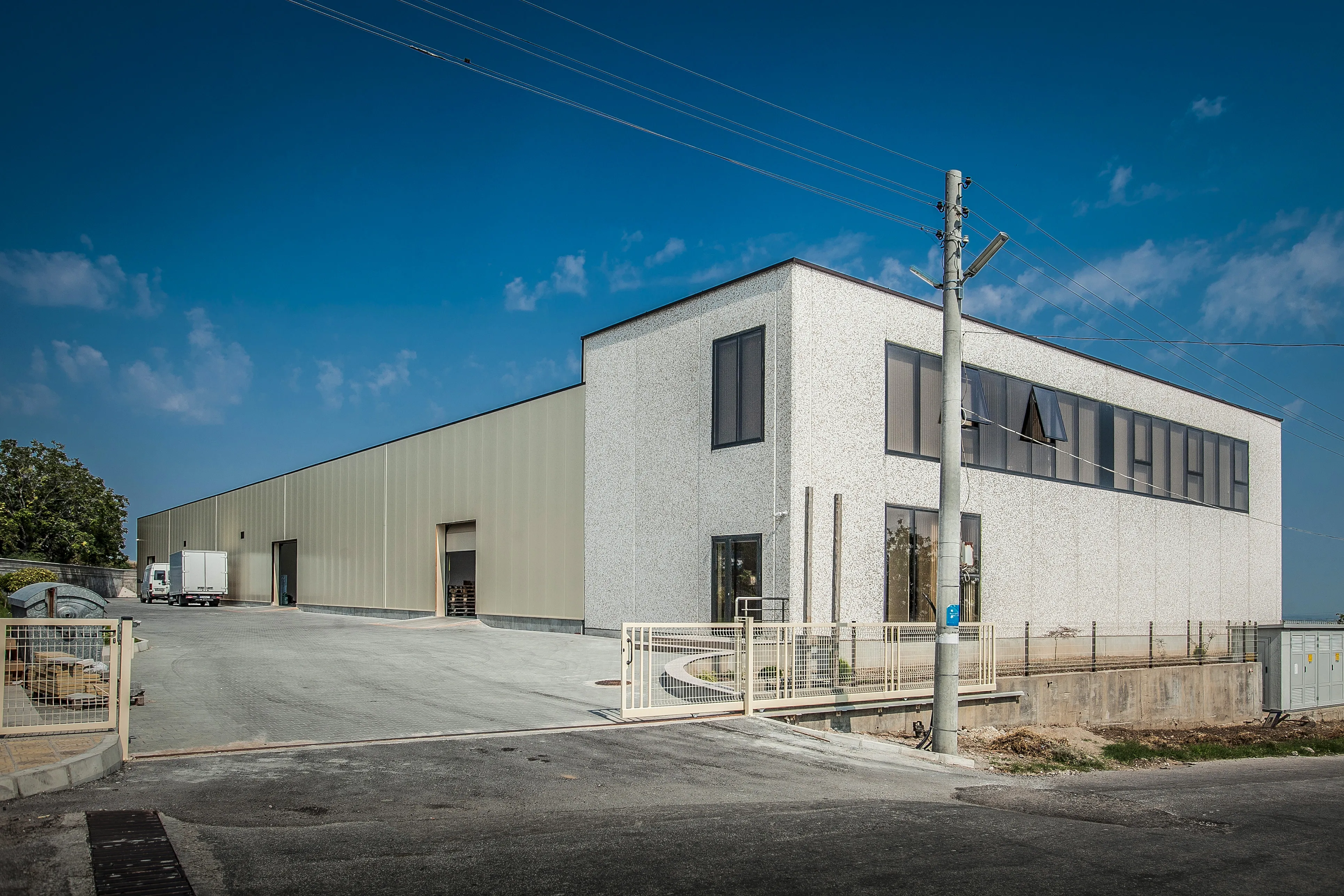 Two-storey industrial building with white textured facade and attached metal warehouse, aerial city view in background