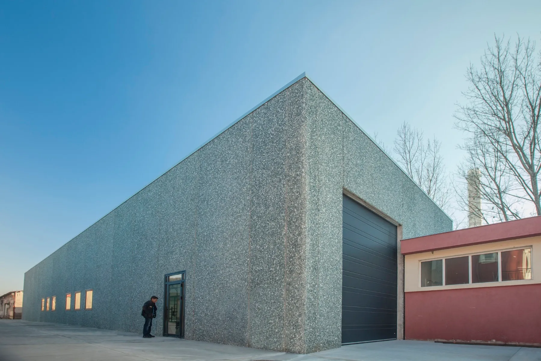 Single-storey industrial warehouse with stone-textured grey facade and large roller shutter door, viewed at dusk
