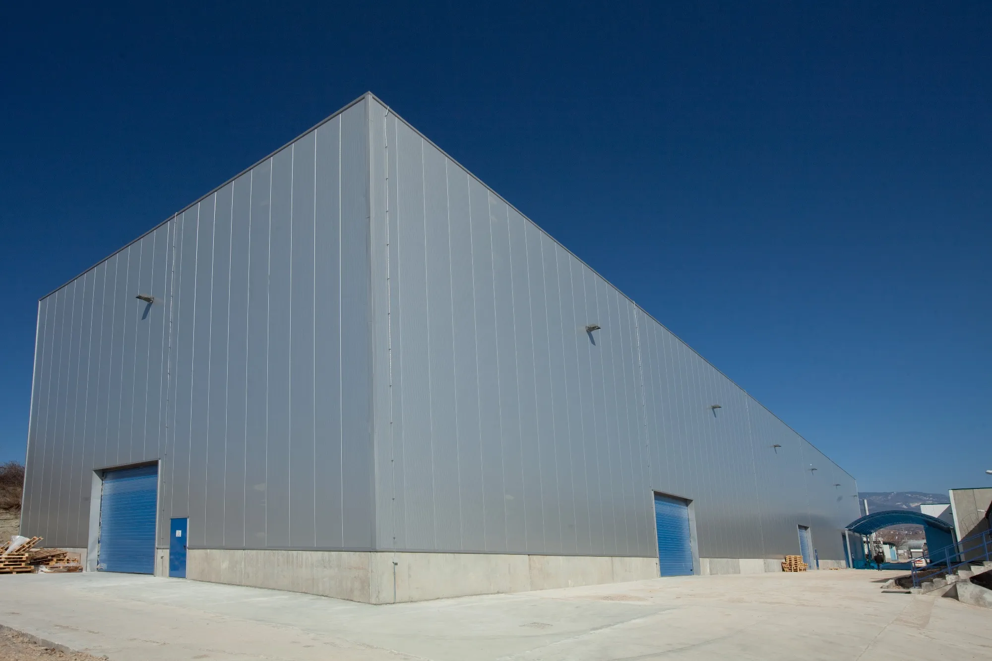 Large grey metal-clad industrial warehouse with two blue roller shutter doors under a clear deep blue sky