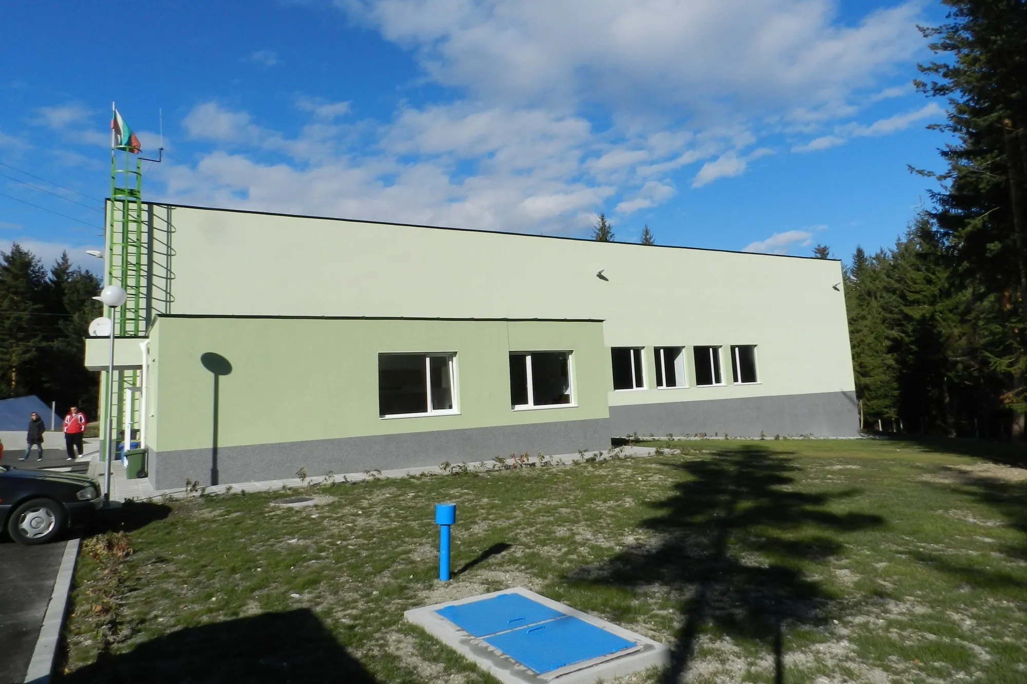 Single-storey public utility building with green facade, metal mast with Bulgarian flag, surrounded by pine forest