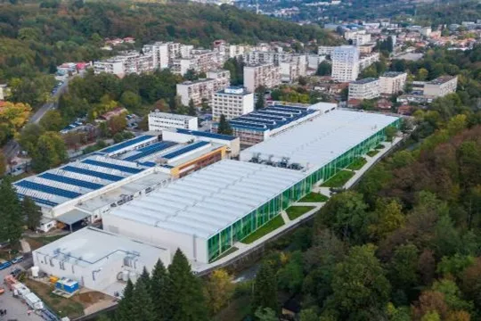 Aerial view of large industrial facility with barrel-vault skylights and green glass facade amid forested city hills