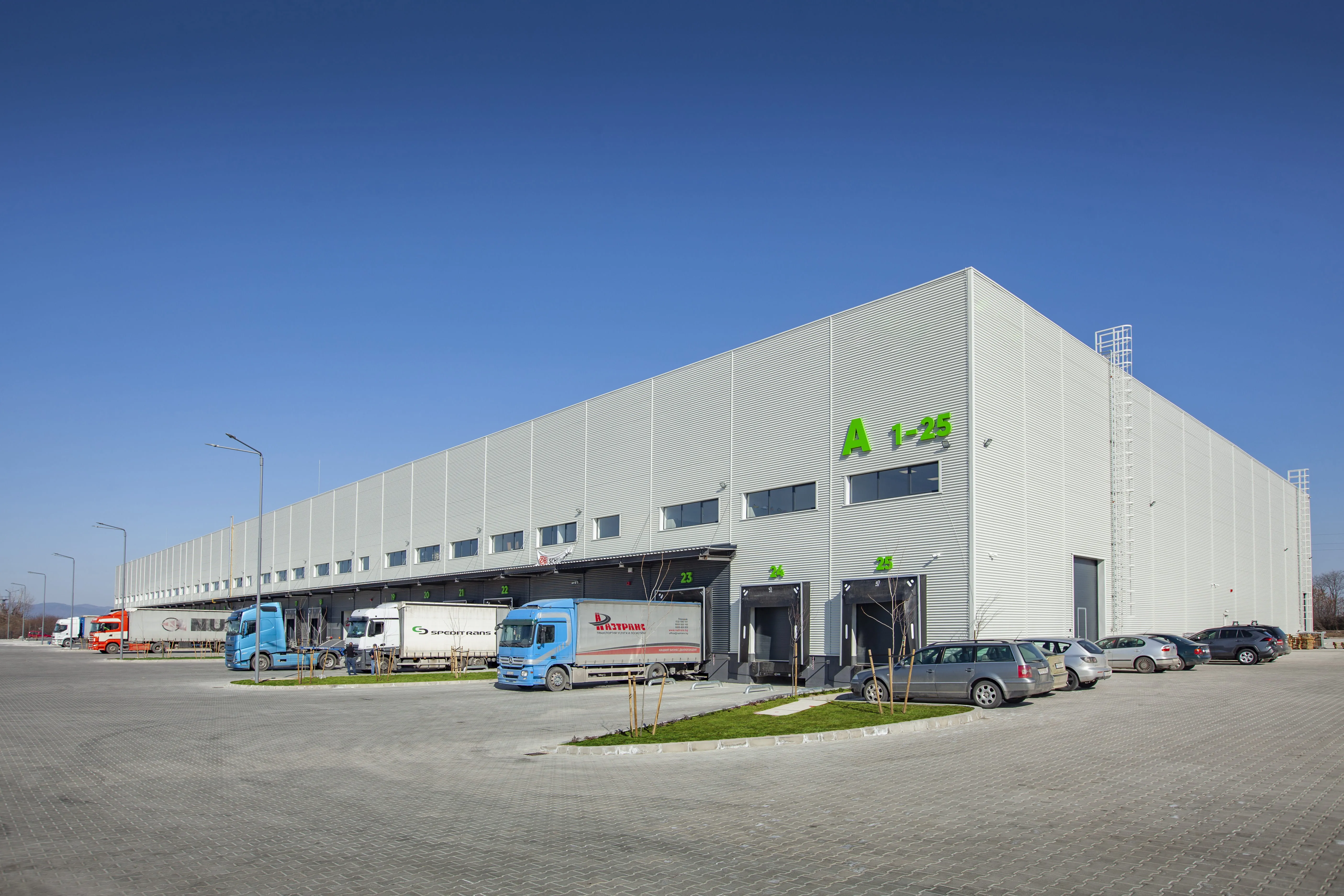 Exterior of a large grey logistics warehouse labeled A 1-25 with multiple loading docks and parked trucks