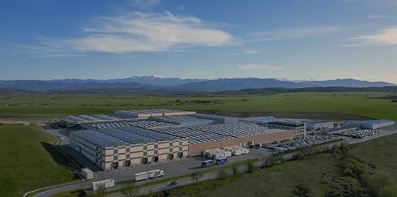 Aerial view of a large warehouse with solar panels on the roof in a green valley with mountains in the background.
