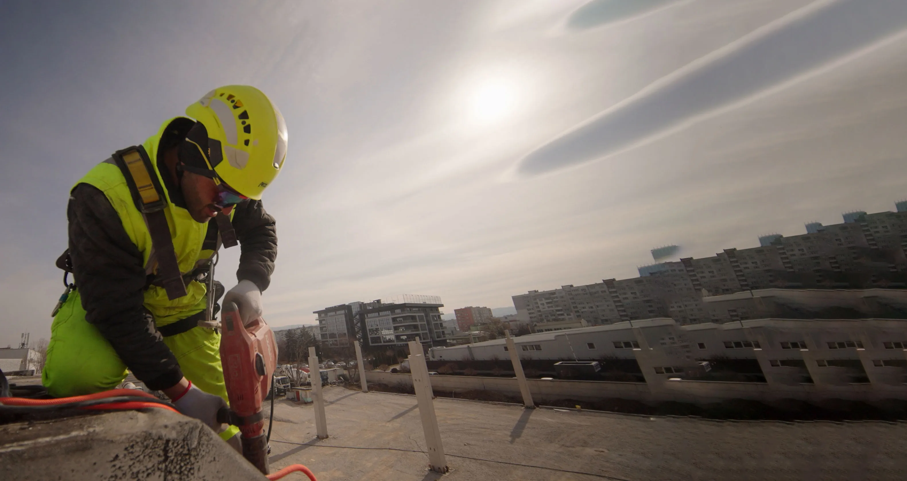 Construction worker in bright safety gear using a power drill on a rooftop with city buildings in the background.
