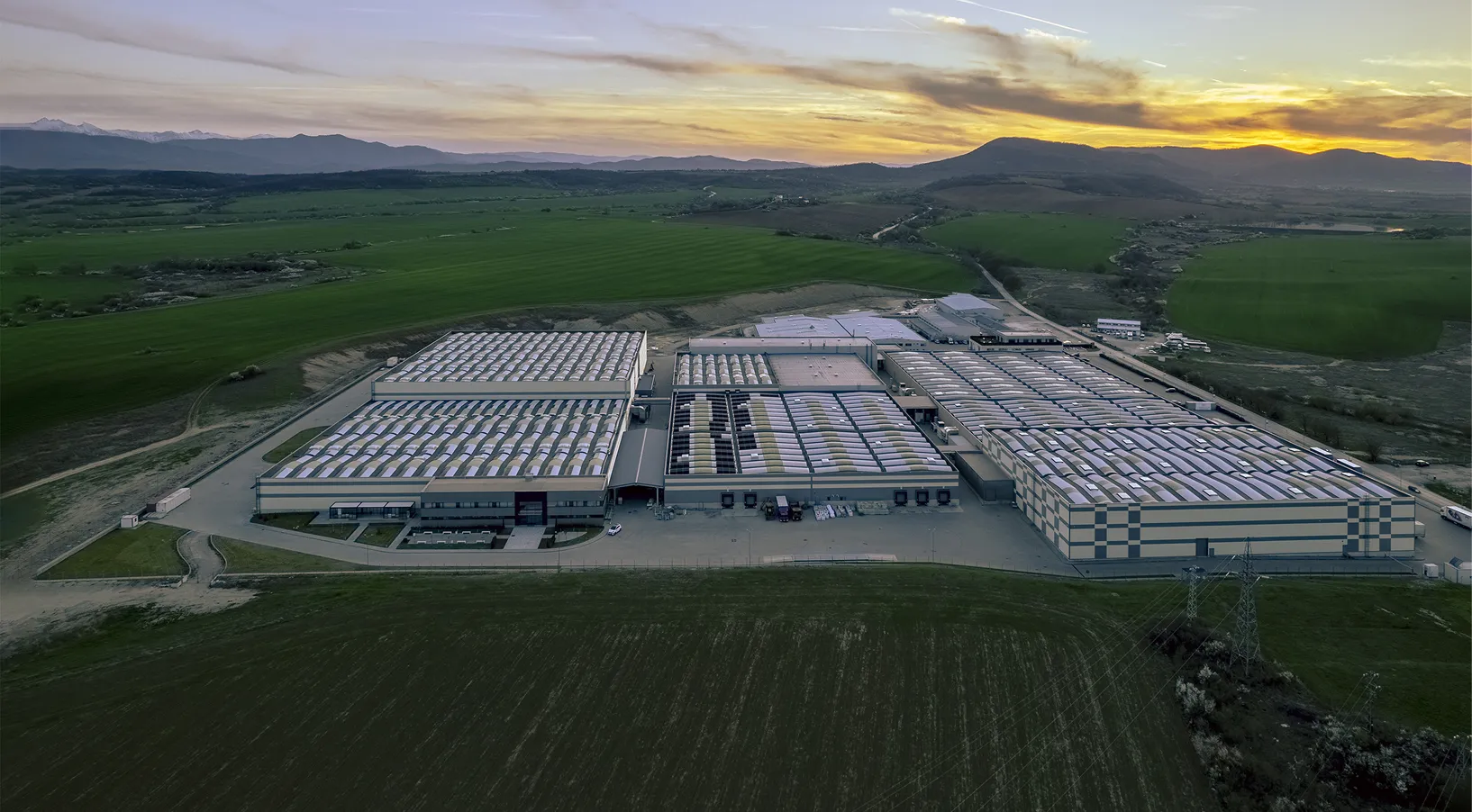 Large industrial complex surrounded by green fields and mountains under a sunset sky.