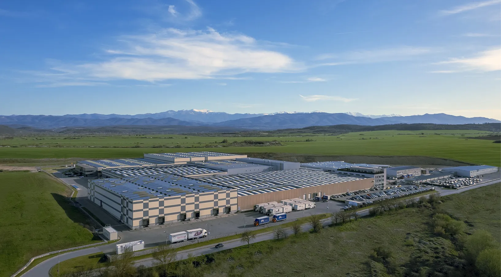 Large industrial warehouse with trucks and cars near green fields and distant mountains under blue sky.