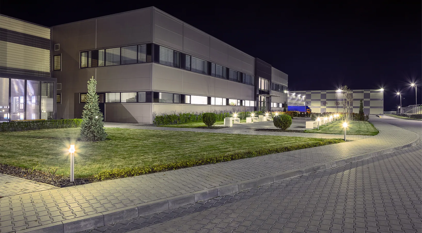 Night view of a modern office building with lit pathways, lawn, and parked blue truck in background.