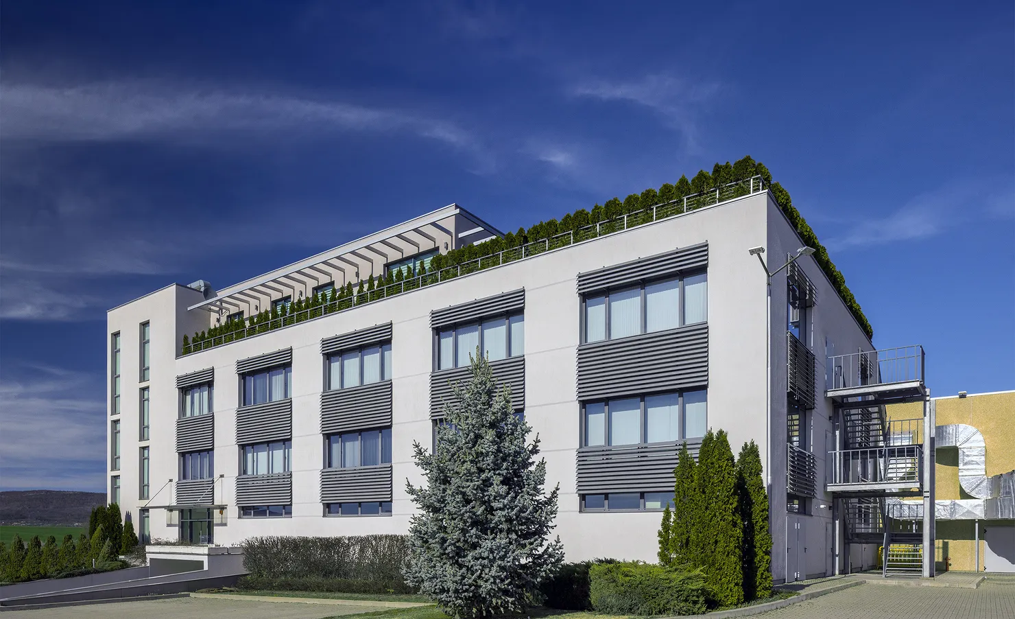 Modern white building with large windows and rooftop greenery under a clear blue sky.
