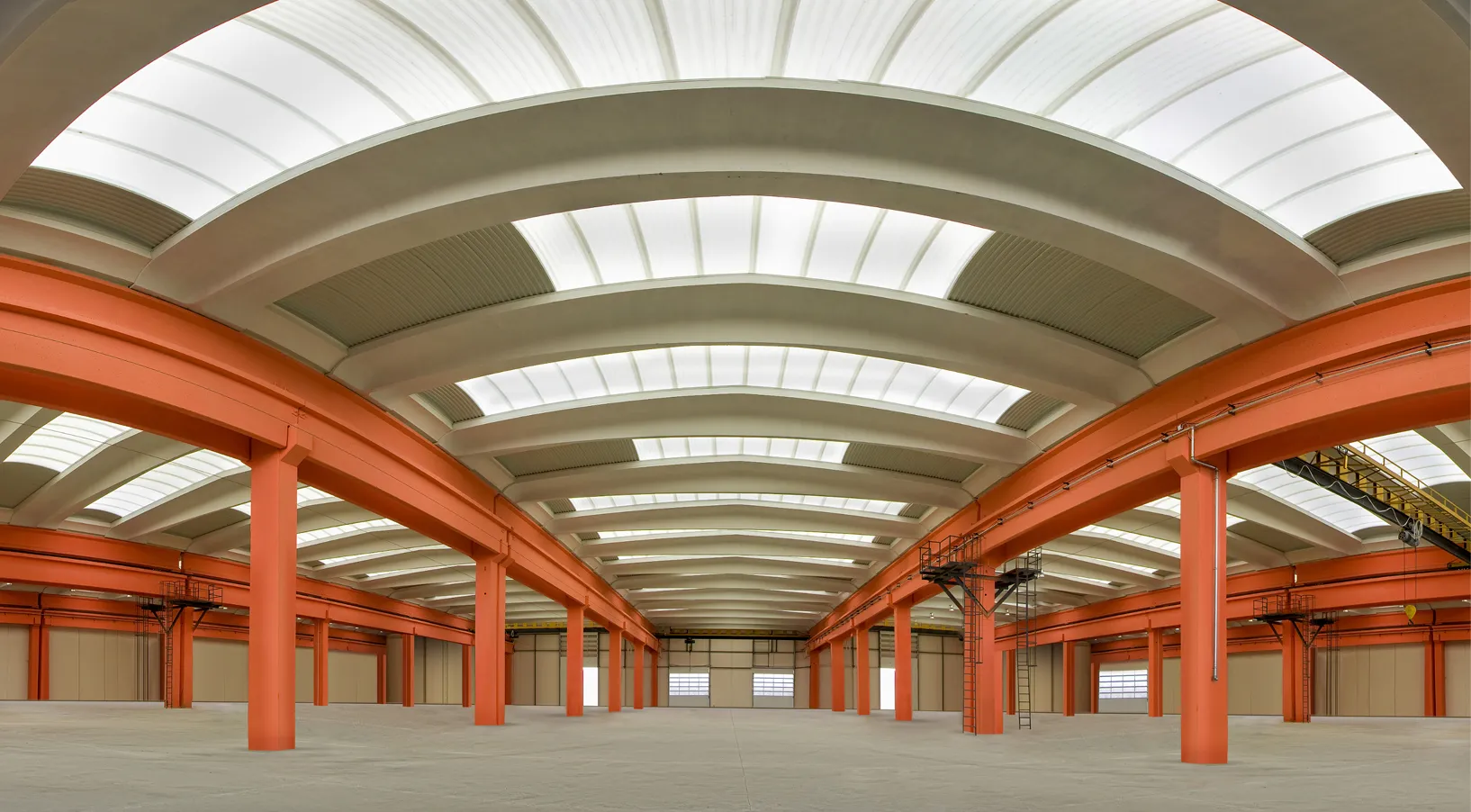 Wide empty warehouse interior with orange steel beams and curved white skylight ceiling.