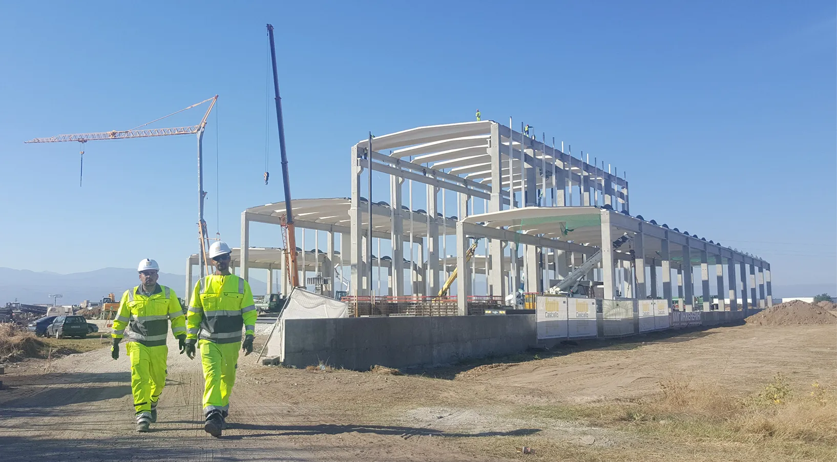 Two construction workers in yellow gear walking near a large building framework under a clear sky.
