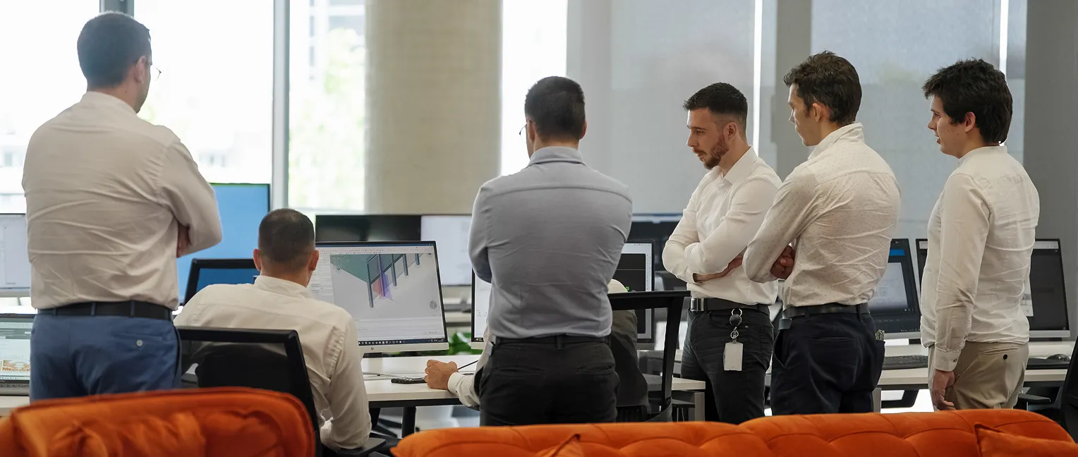Six men in office attire gathered around desks with computer monitors displaying technical drawings.