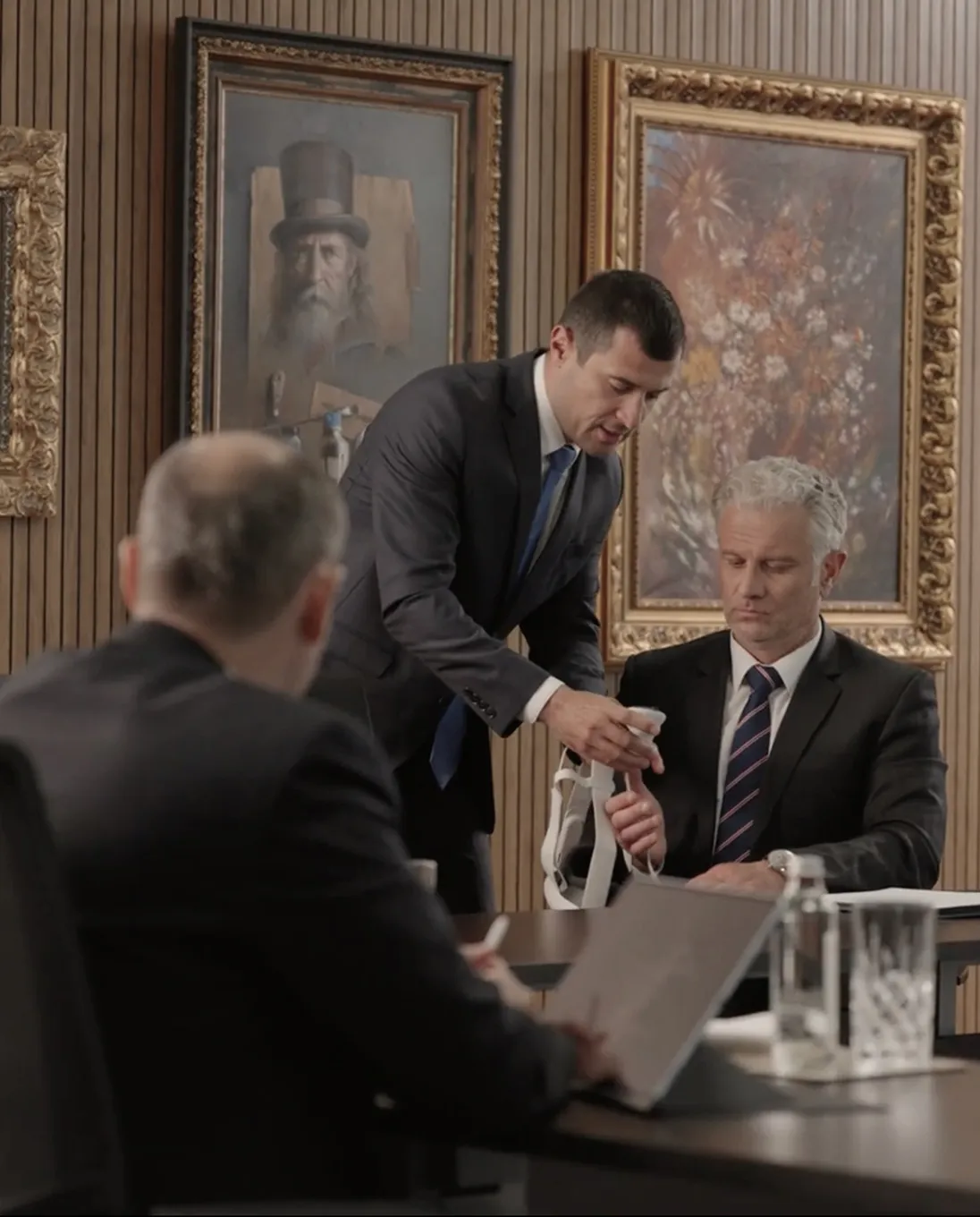 Two men in suits at a table, one standing and handing a white object to a seated man with gray hair.