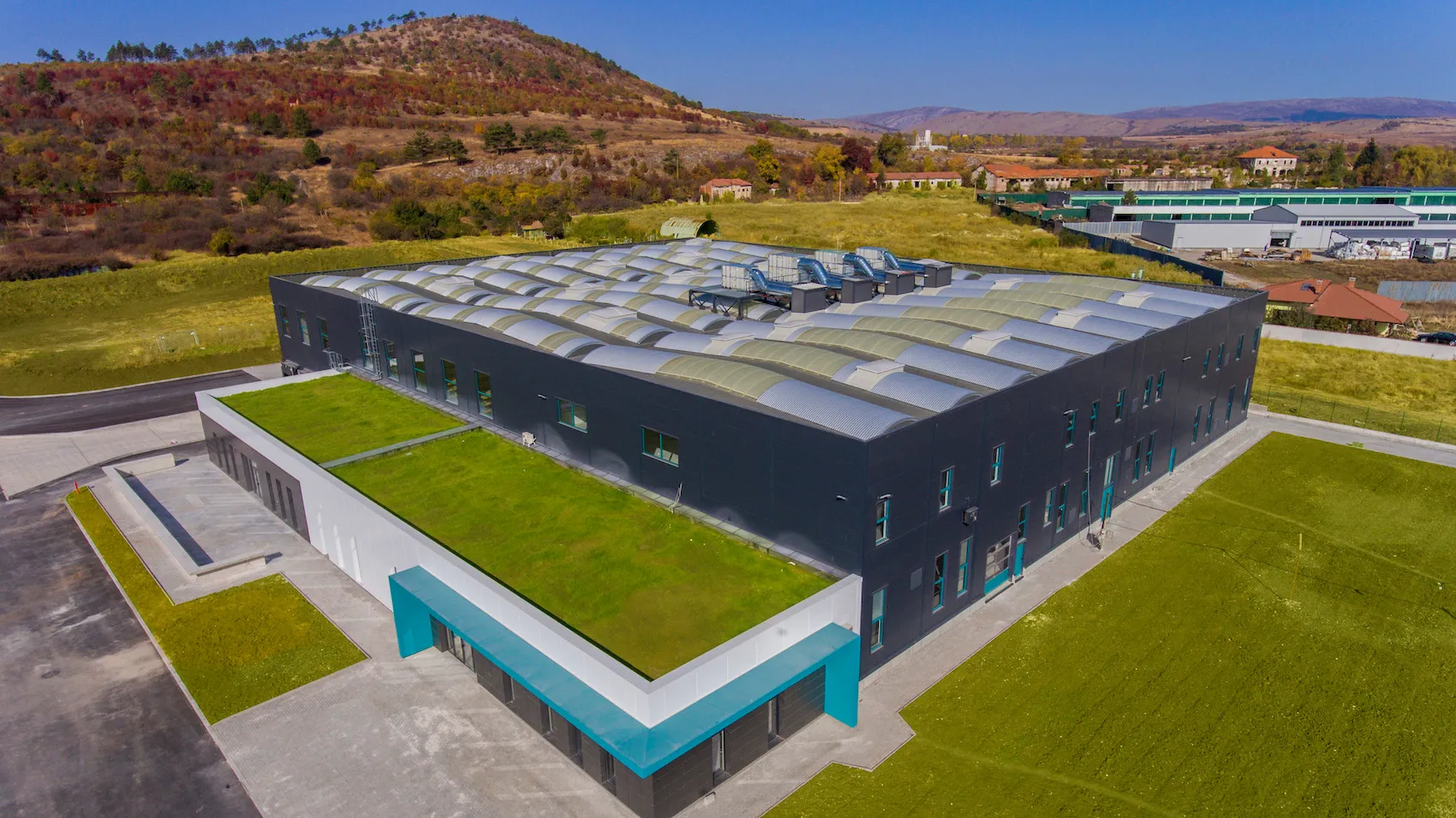 Large modern industrial building with green rooftop, surrounded by grass and hills in the background.