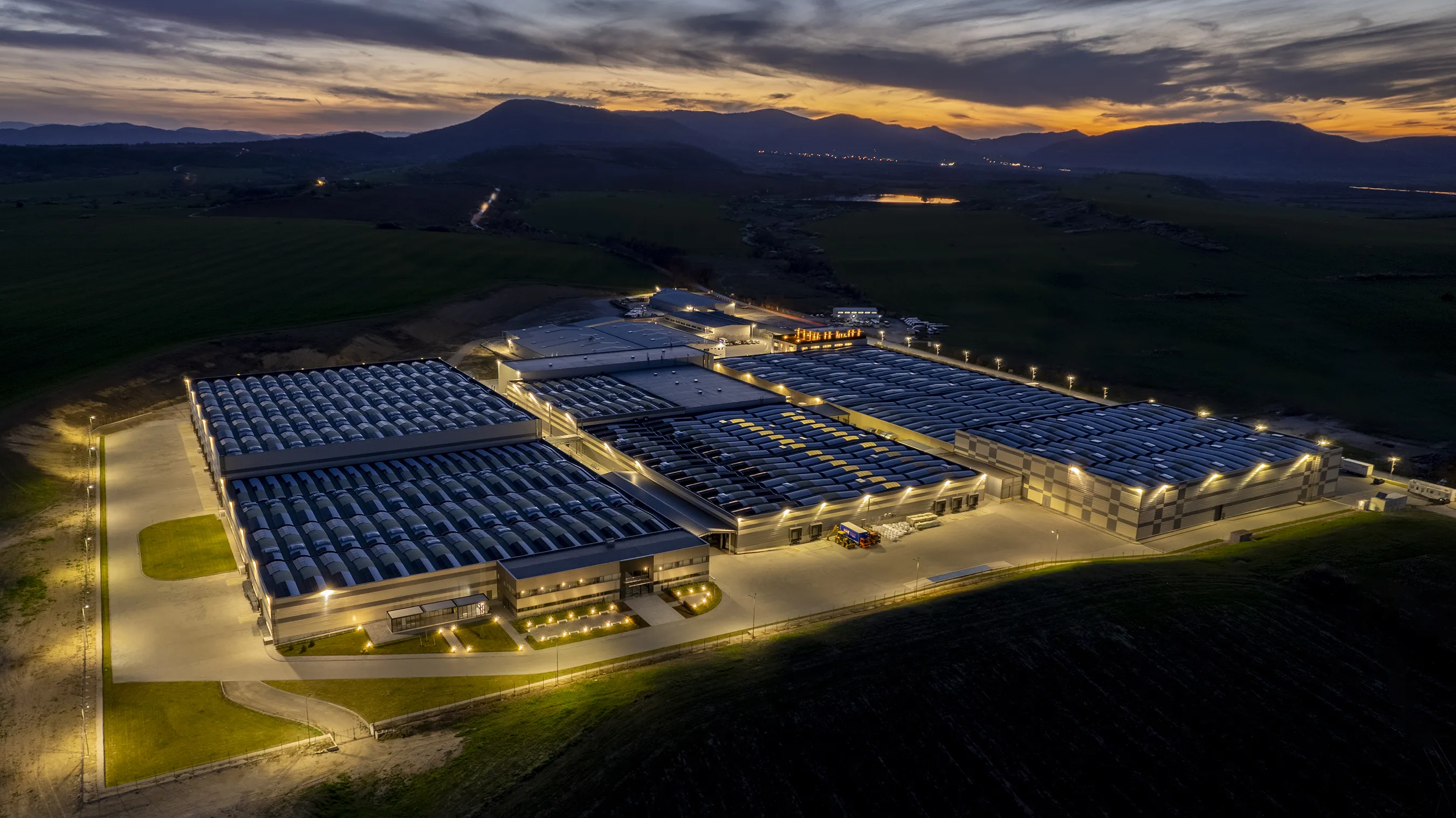 Aerial view of an illuminated industrial factory complex at dusk with surrounding fields and distant mountains.