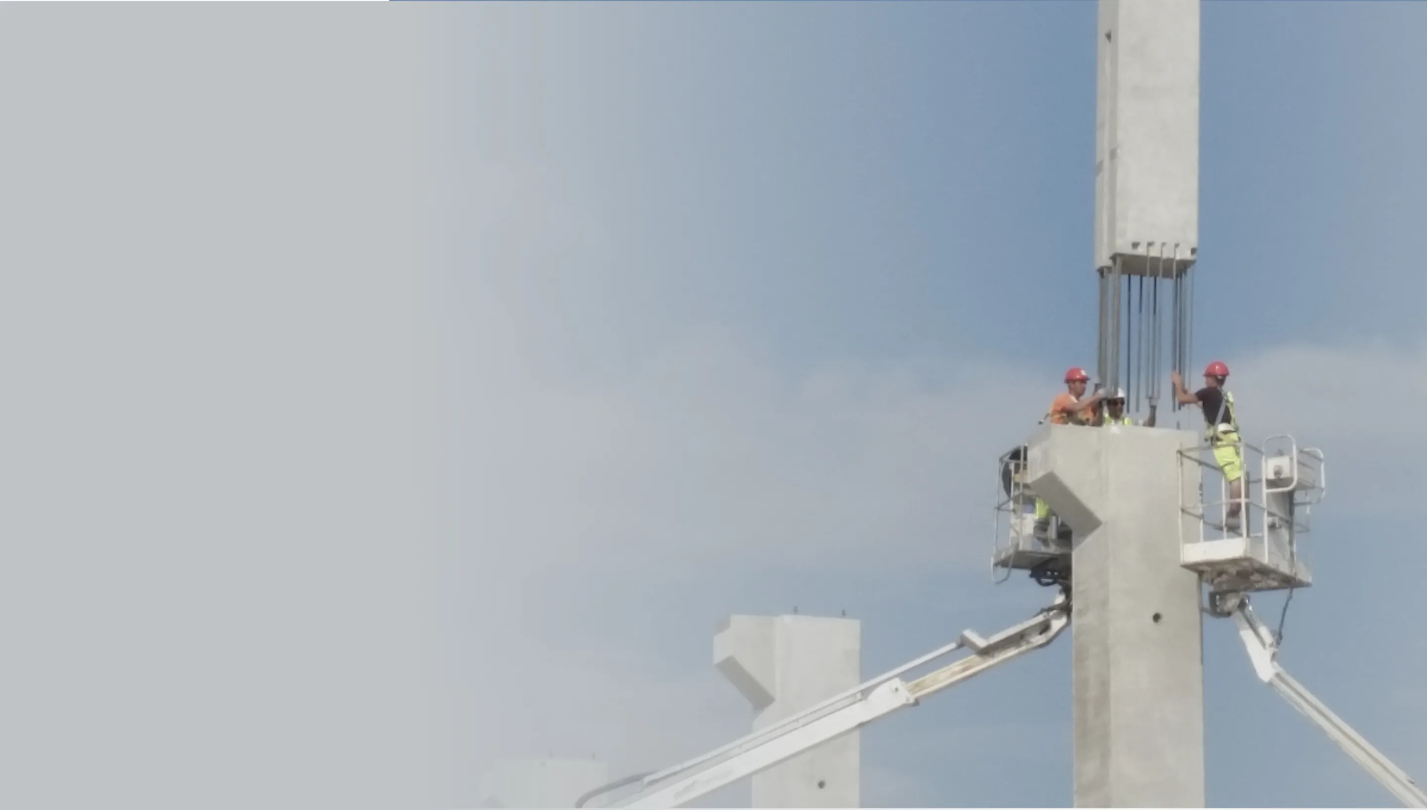 Construction workers wearing helmets and safety vests working on tall concrete pillars with a blue sky background.